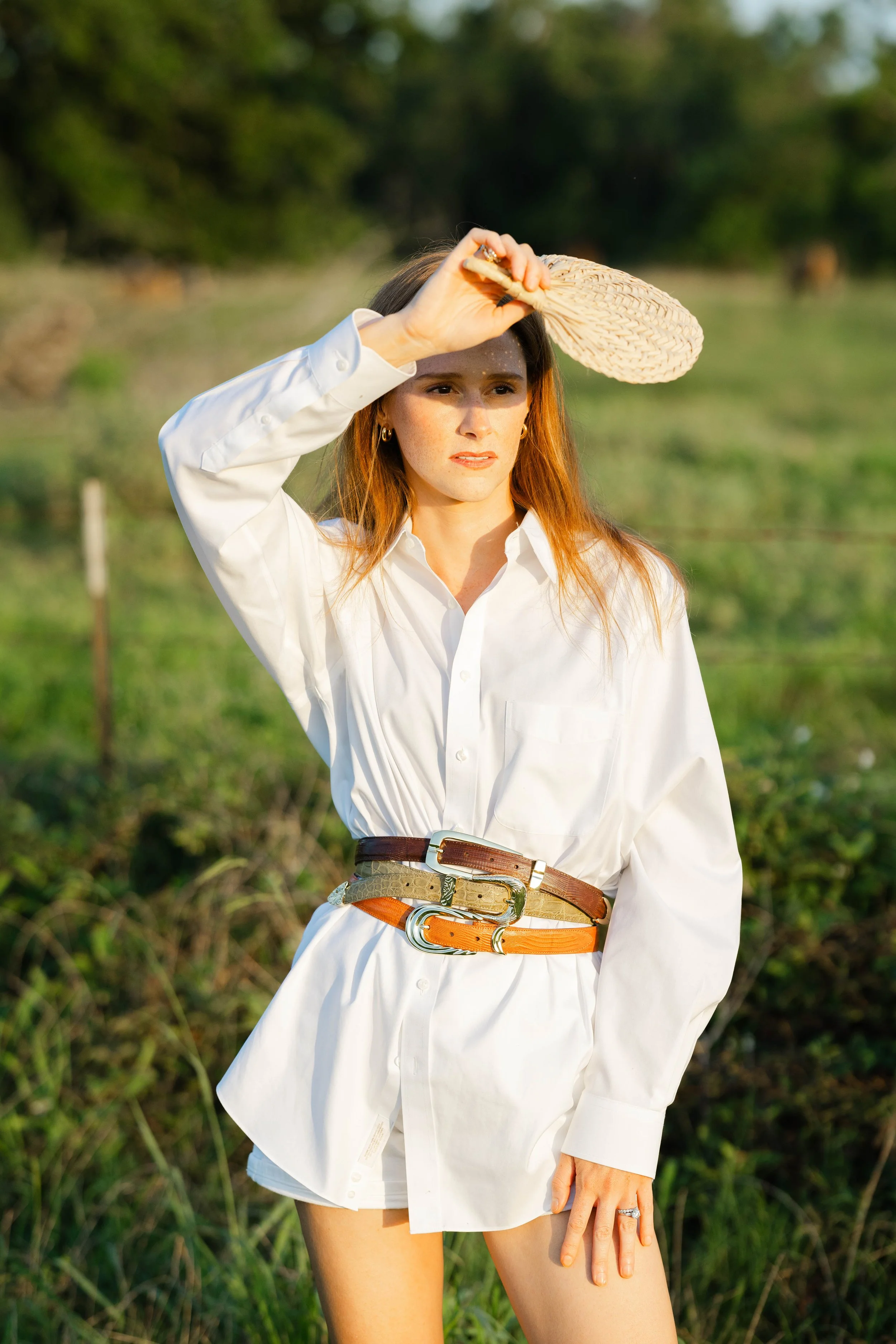 A woman with long brown hair wearing a white button-down shirt, a wide multicolored belt, and holding a woven straw hat, standing outdoors in a grassy field with trees in the background.