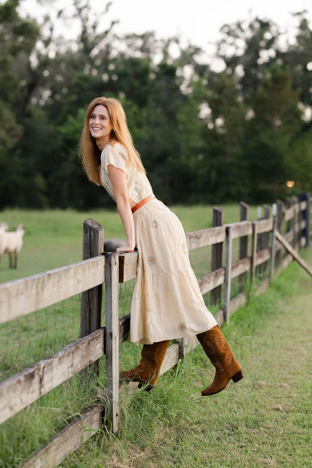 A young woman with long red hair smiling and leaning over a wooden fence in a rural field with trees in the background. She is wearing a cream-colored dress with embroidered details and brown cowboy boots.