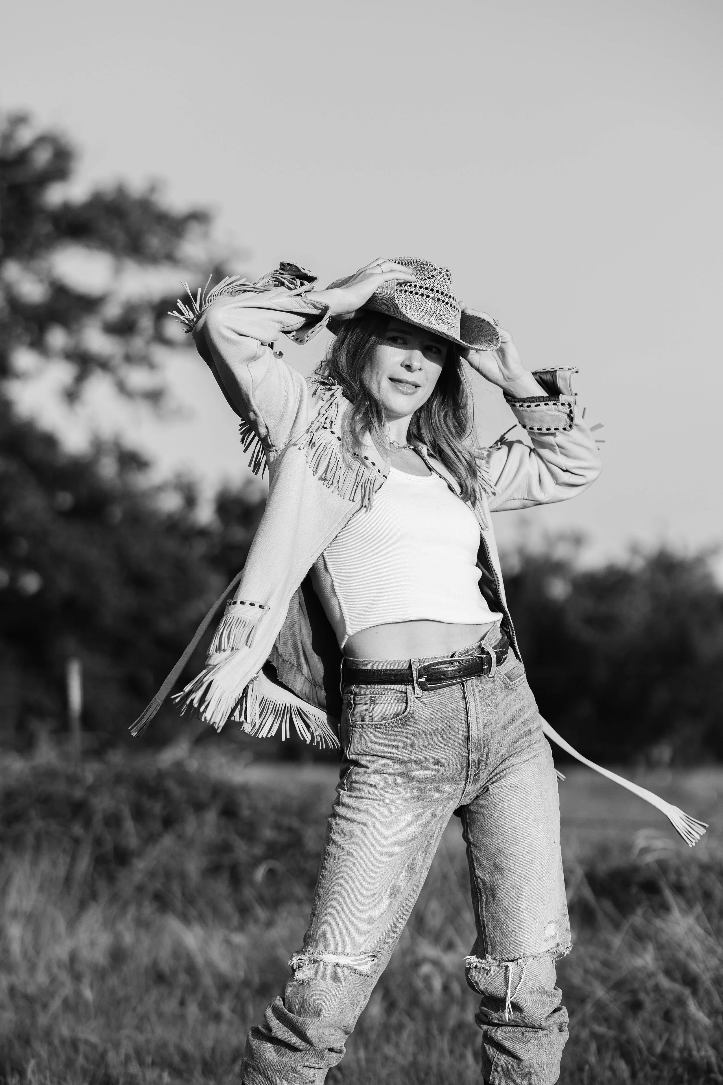 A woman wearing a fringed jacket, jeans, and a wide-brimmed woven hat, standing outdoors in a rural setting with blurred trees in the background, looking at the camera.
