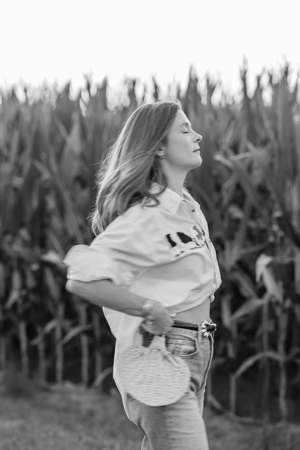 A woman with long hair is walking outdoors in front of a cornfield, with her eyes closed and a peaceful expression.