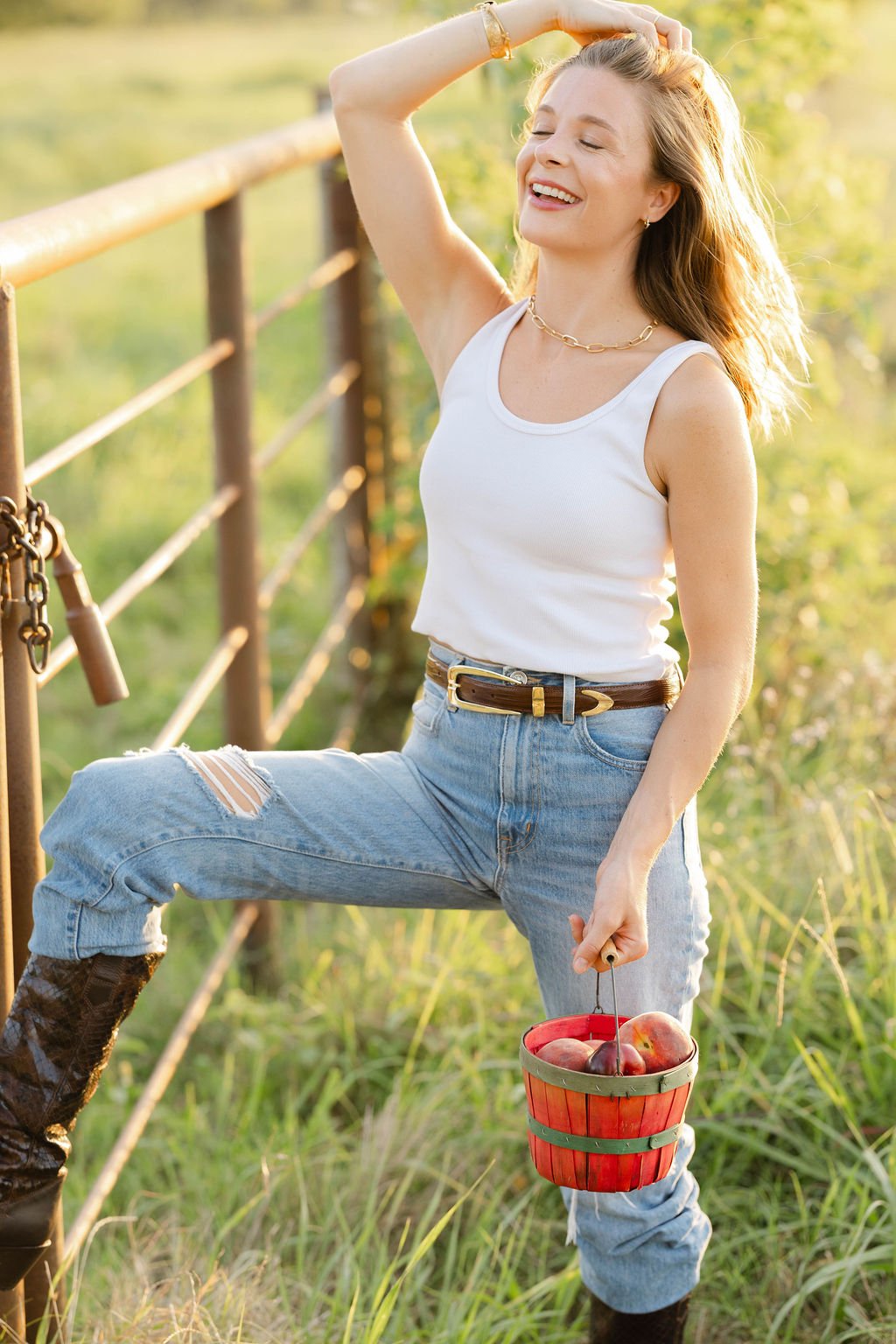 A woman with blonde hair, wearing a white tank top, ripped jeans, and black boots, stands outdoors on a grassy field with greenery in the background. She has a chocolate brown leather belt on with a gold buckle.