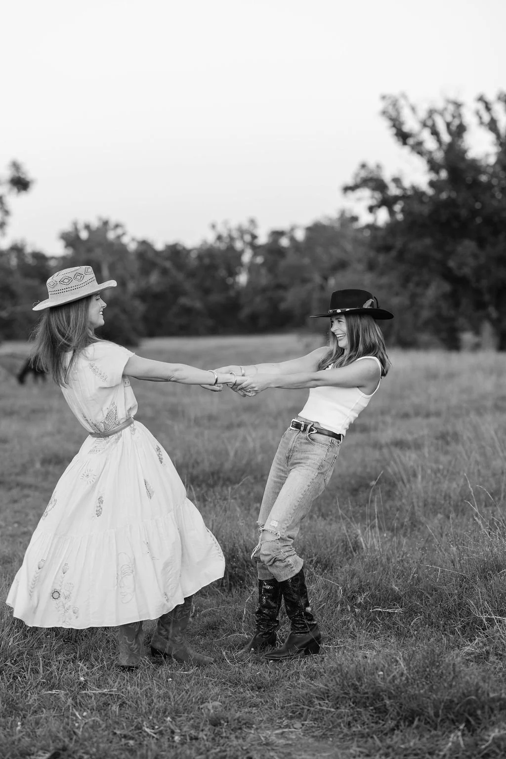 Two women wearing cowboy hats and casual clothing holding hands and dancing in a grassy field with trees in the background.