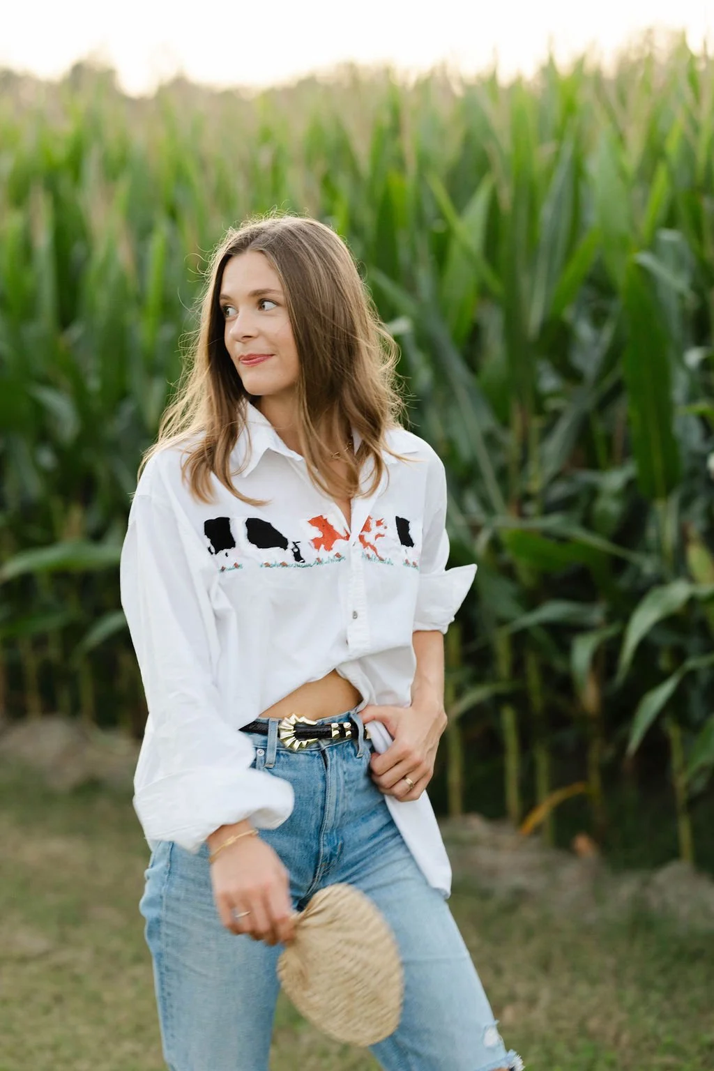 A young woman with brown hair wearing a white embroidered shirt and blue jeans, holding a straw hat, standing in front of a cornfield.