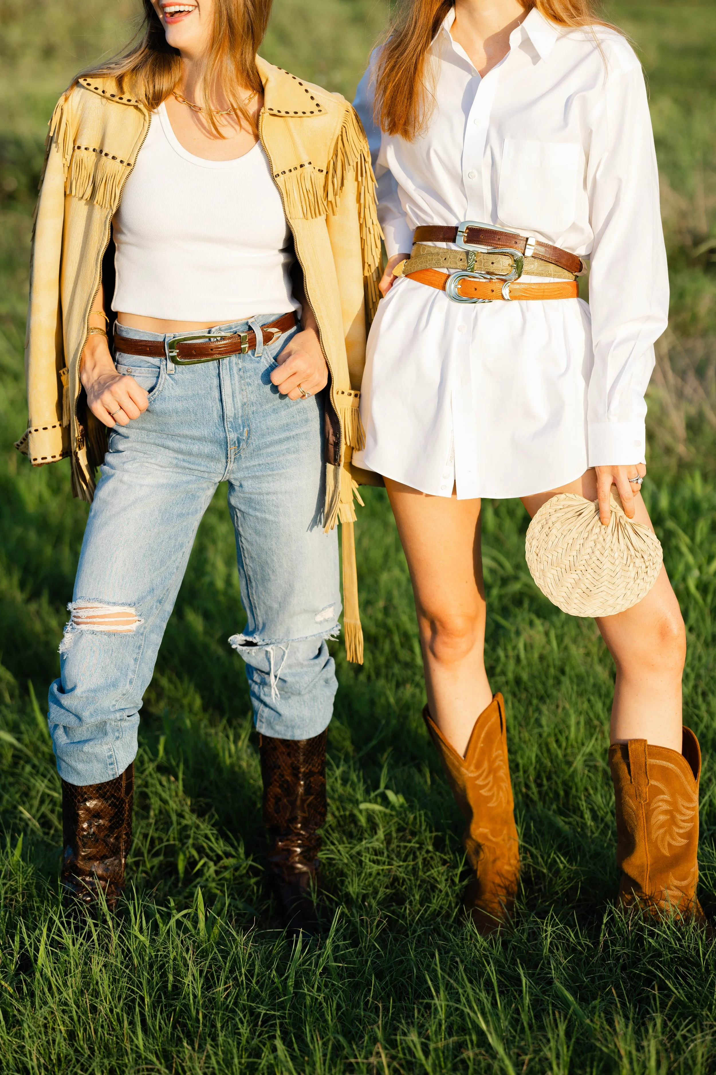 Two women standing in a grassy field, dressed in western-style clothing with cowboy boots, belts, and hats. One wears ripped jeans and a fringed jacket, the other a white shirt dress, both smiling.