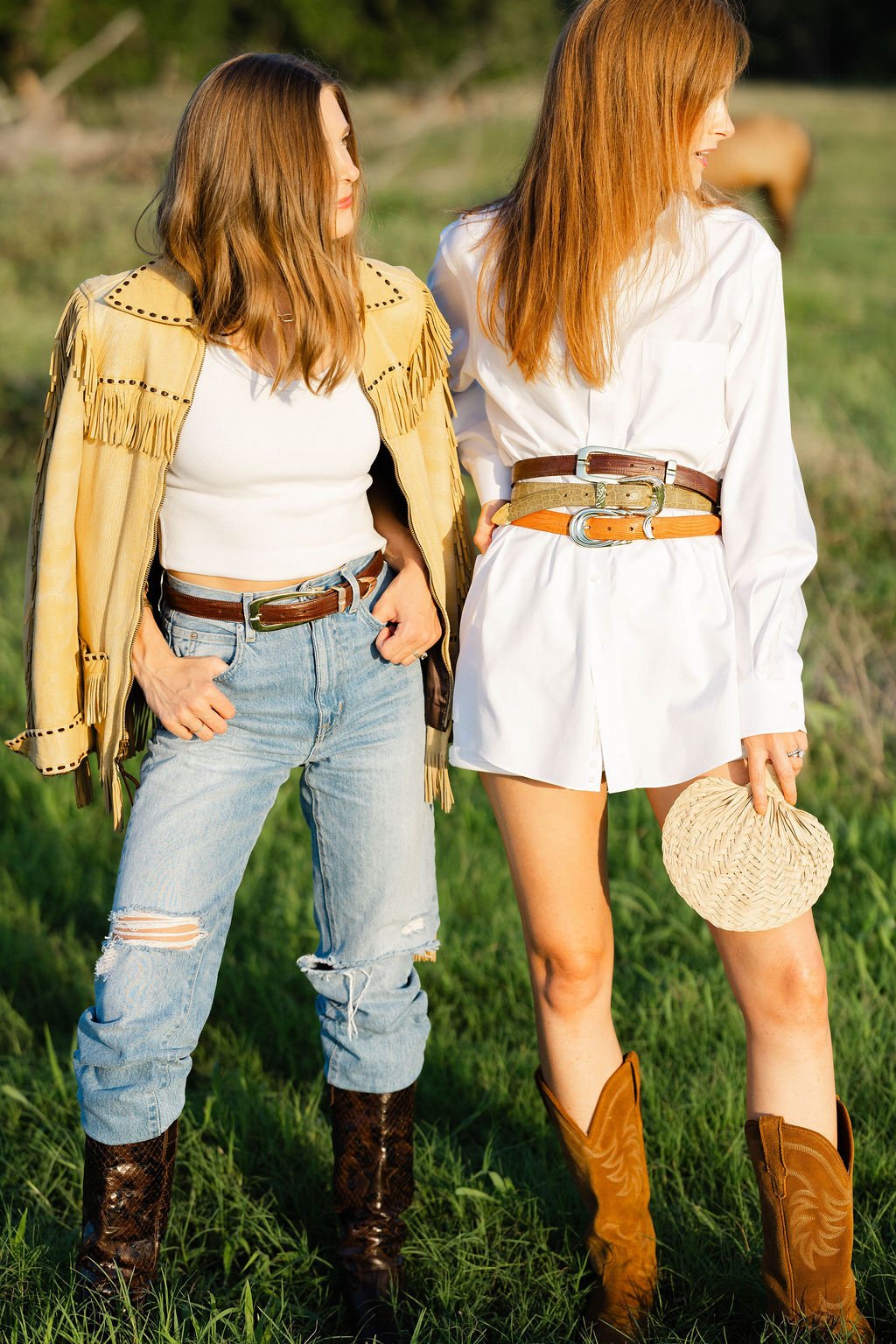 Two women with red hair, dressed in Western attire, standing outdoors in a grassy field, with one holding a straw hat.