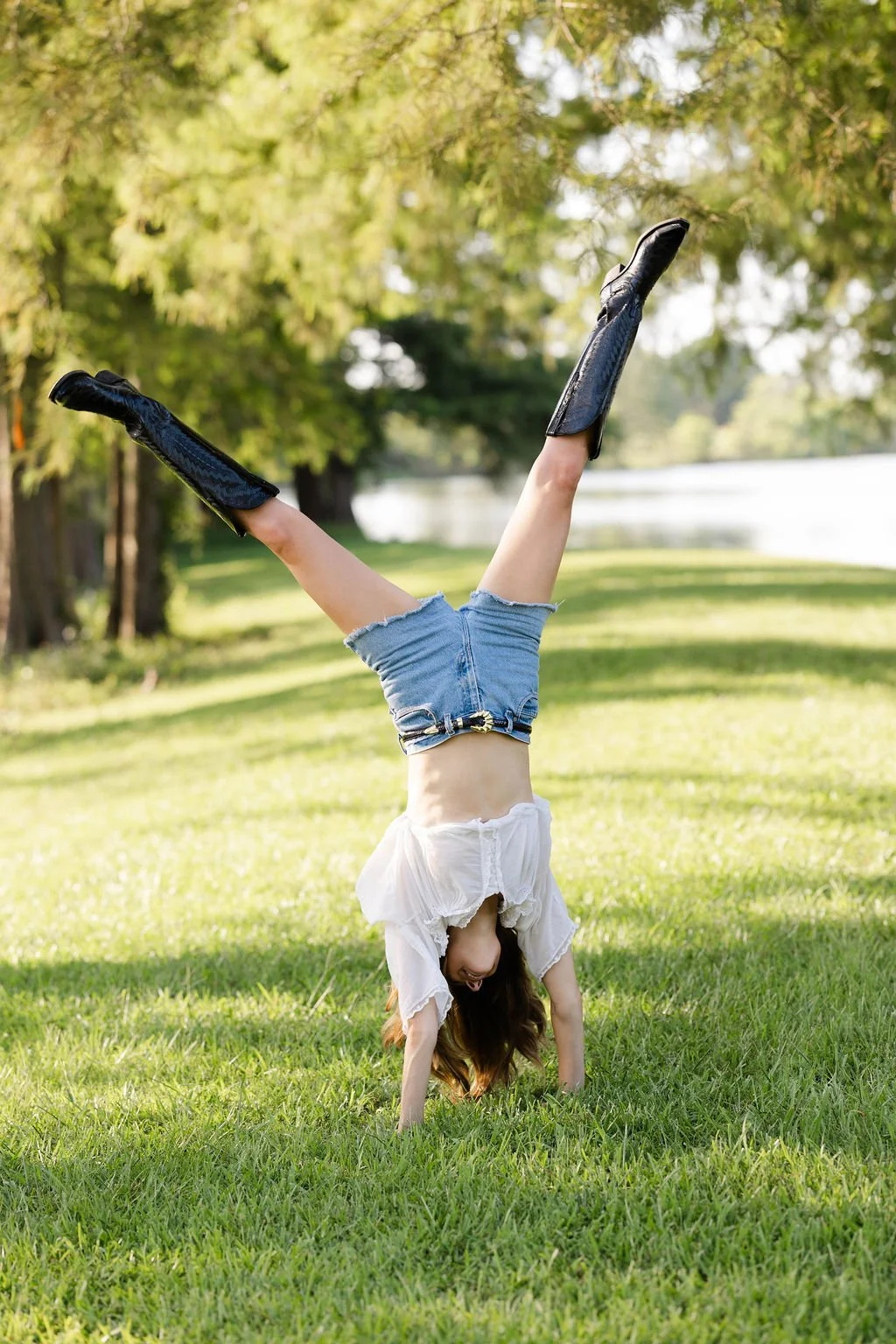 A woman performing a handstand outdoors on a grassy field near a lake, wearing denim shorts, a white blouse, and black knee-high boots.