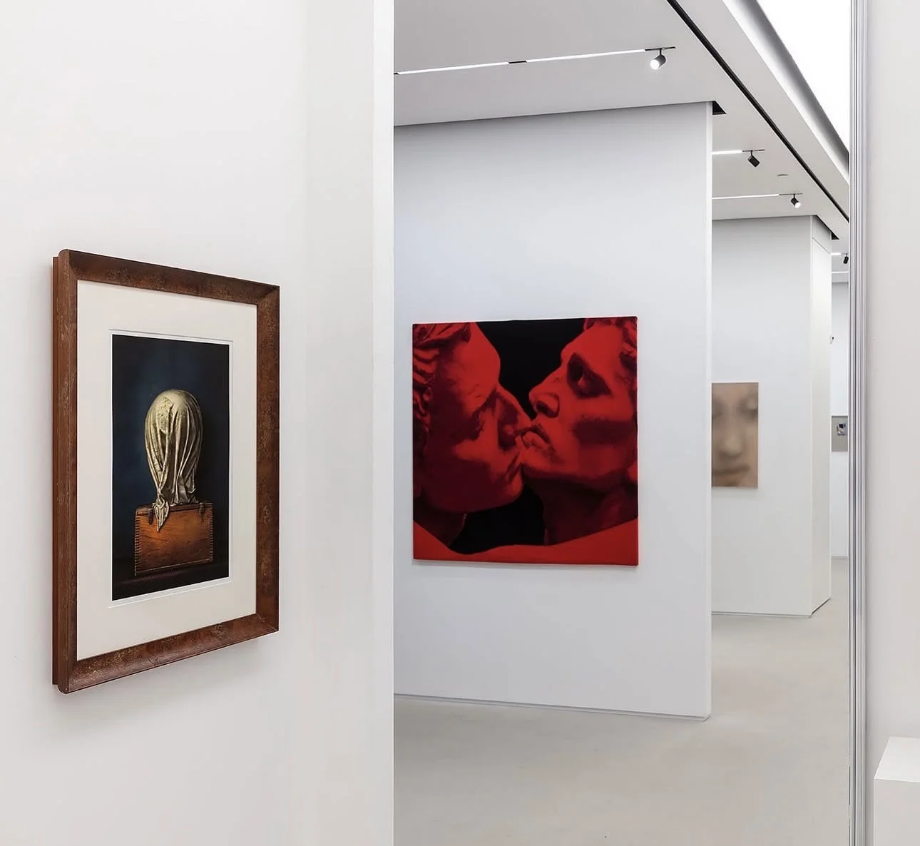 Interior of an art gallery with paintings on white walls, including a framed painting of a figure with a gold head and a red painting of two faces close together.