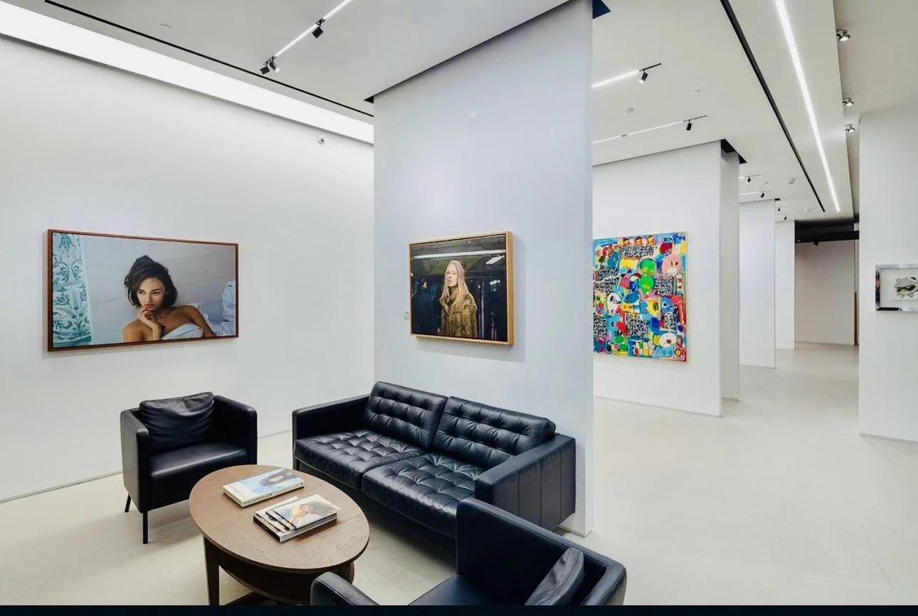 Modern art gallery with white walls, black leather sofas, and colorful paintings, including portrait photographs and abstract art.