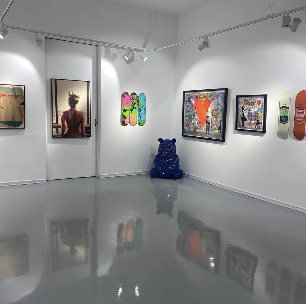 Contemporary art gallery with various colorful paintings on white walls, a glossy gray floor, a blue bear sculpture, and skateboard art on the wall.