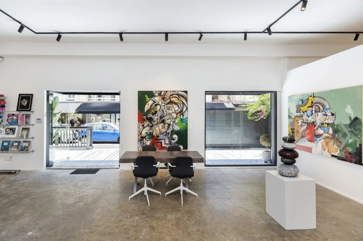 An art gallery with two large abstract paintings on the walls, a table with four chairs, a sculpture of stacked stones on a white pedestal, and a magazine rack on the left wall.
