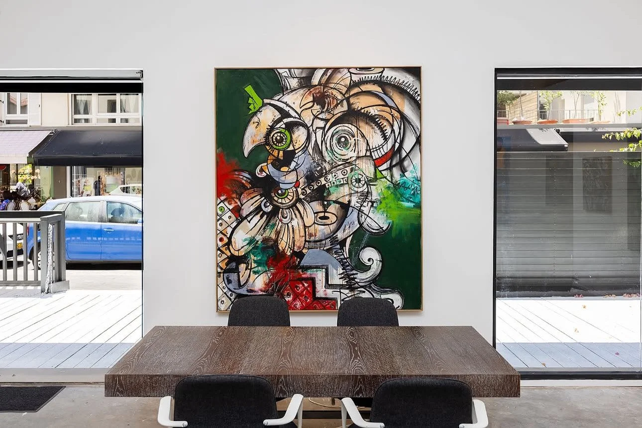 A modern dining area with a wooden table and three black chairs, featuring a colorful abstract mural of a bird on the white wall behind. Large windows on either side reveal a street view with a blue car parked outside.