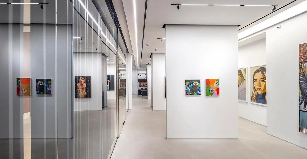 Gallery interior with colorful abstract paintings on the walls and a reflective glass wall on the left side.