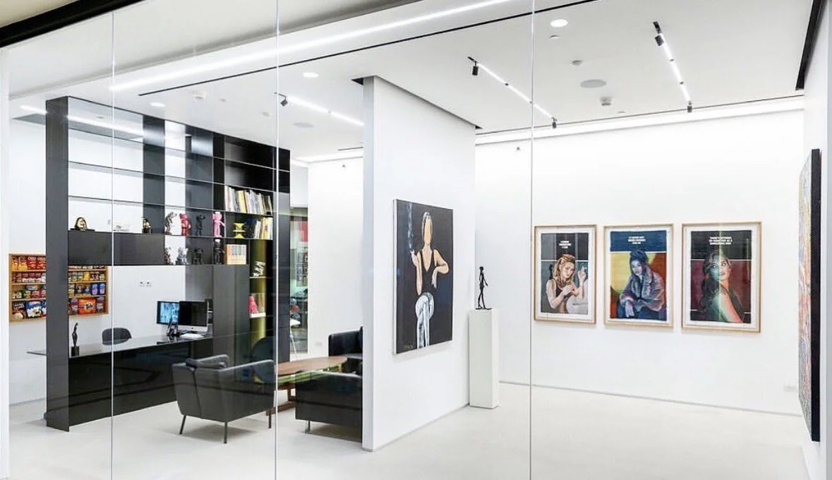 Photo of an art gallery with paintings on white walls, a desk with chairs, a computer, and shelves with books and decorative objects.