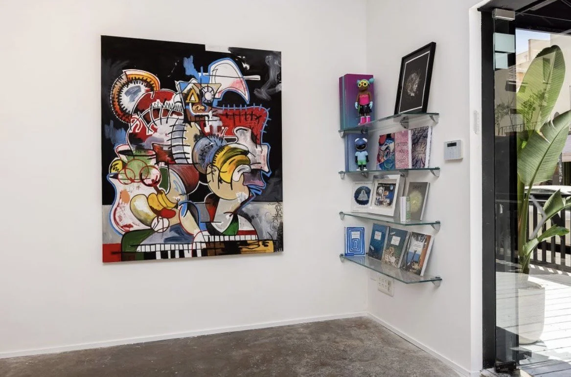Gallery wall with colorful abstract artwork, a shelf with art figures and books, and a glass door leading outside with a potted plant nearby.