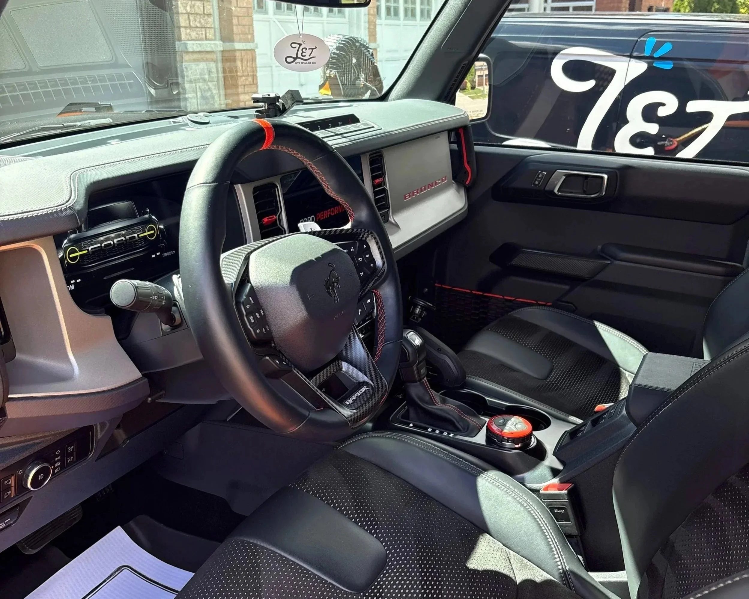 Interior of a Ford Bronco SUV with black and gray seats, a dashboard with various controls, a steering wheel with a flag pattern, and a center console with a drive mode selector and gear shifter.