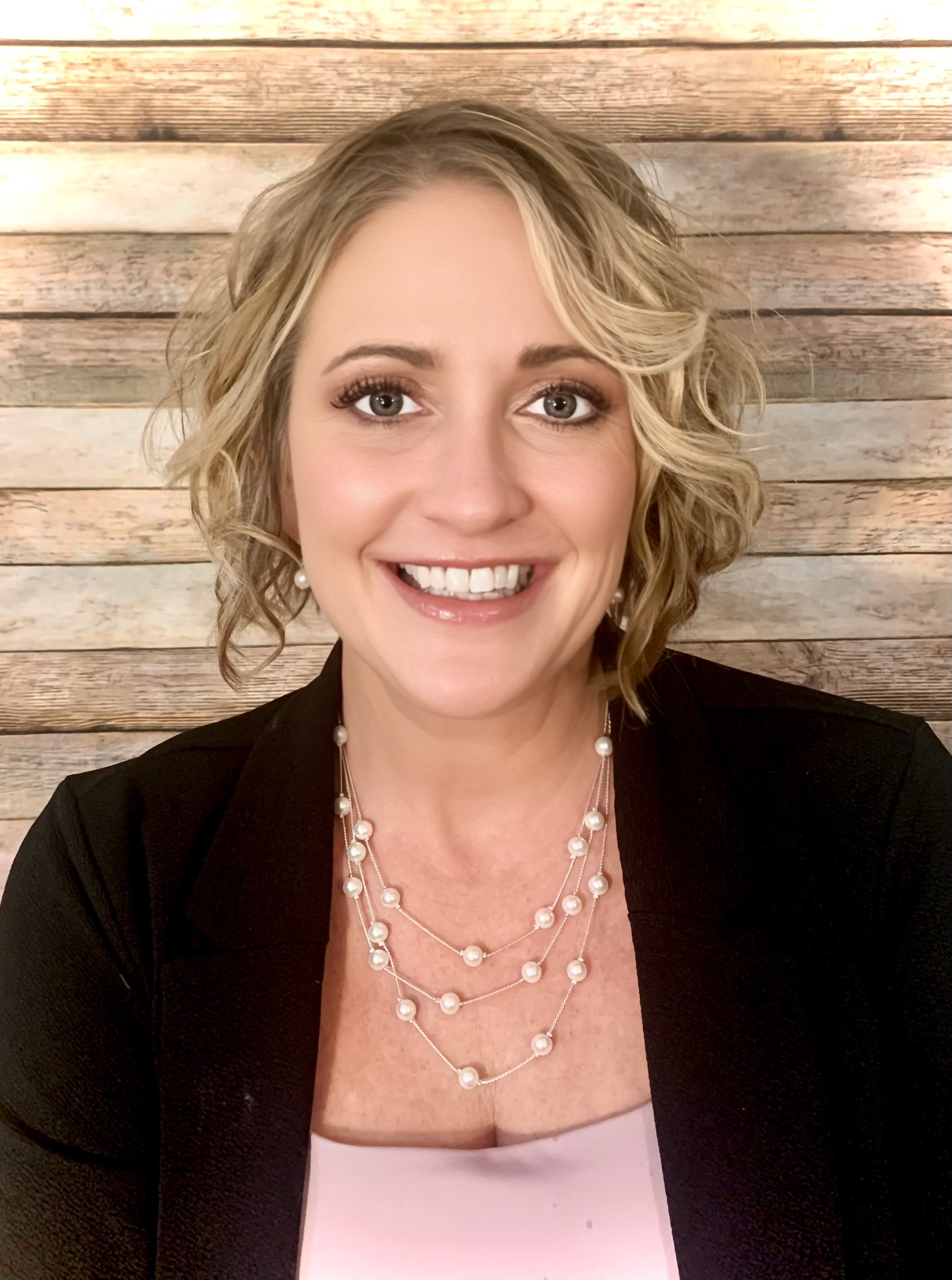 A woman with curly blonde hair smiling, wearing a black blazer and layered pearl necklaces, set against a wooden plank background.