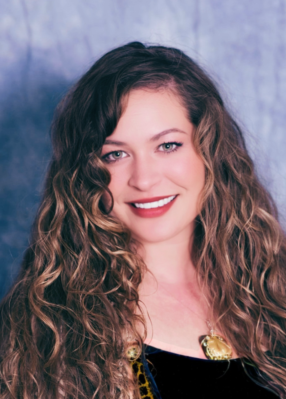 Close-up portrait of a woman with long, curly brown hair smiling at the camera, wearing a black top and gold jewelry, against a blue textured background.