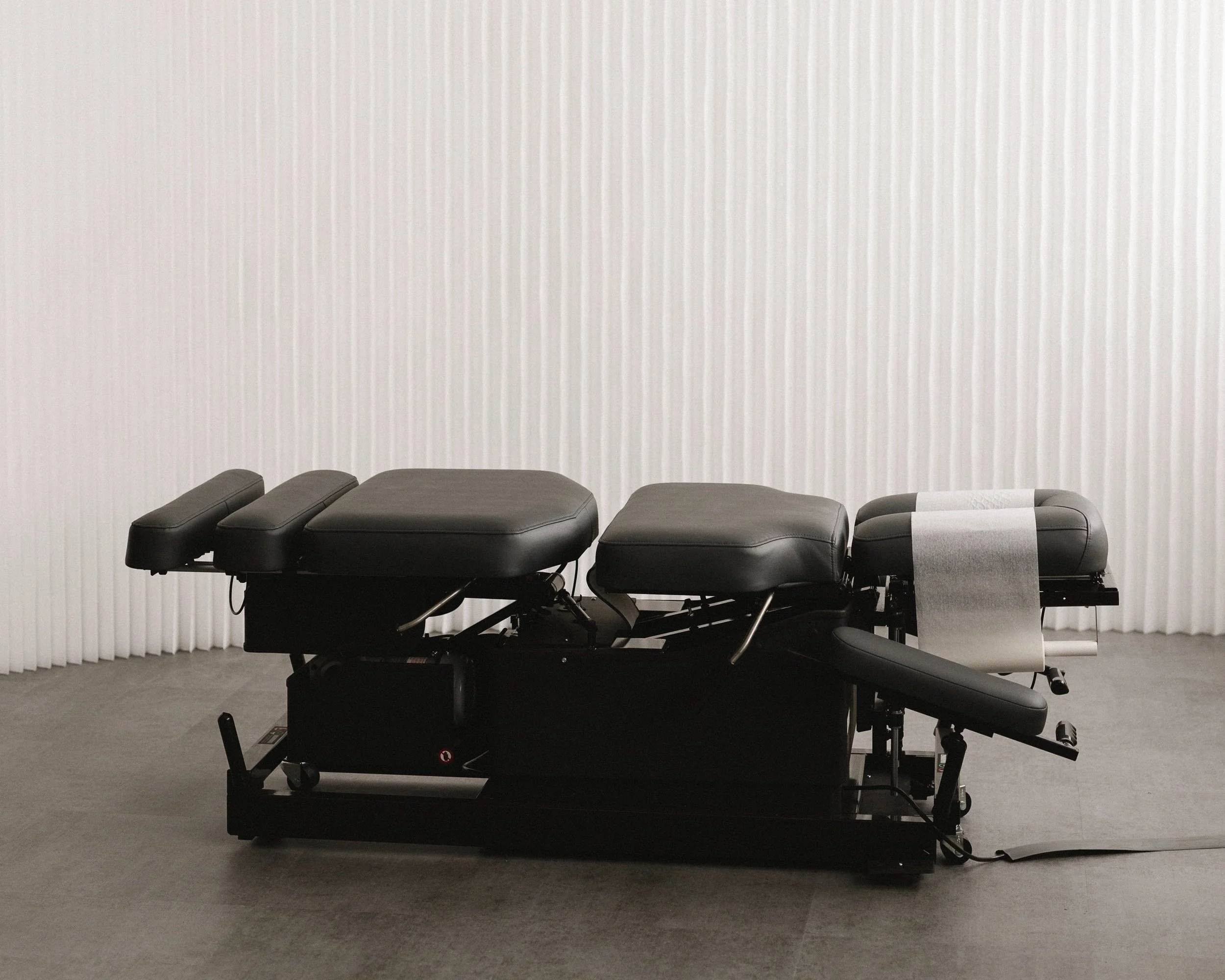 Black medical examination table with adjustable sections in front of a white textured wall.