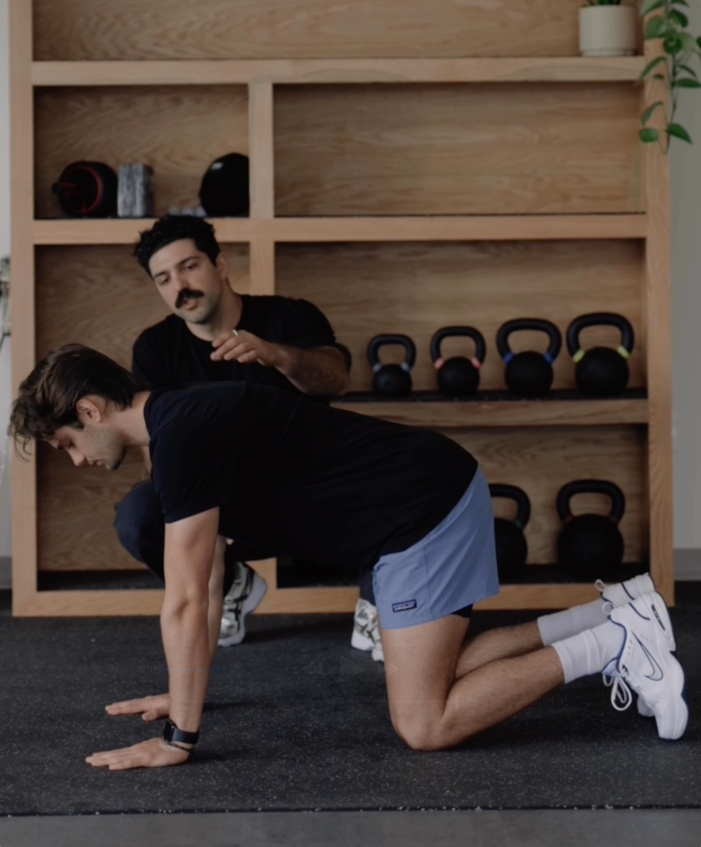 A man in black shirt demonstrating a plank exercise on hands and knees with a trainer instructing him, in a gym with kettlebells on shelves in the background.