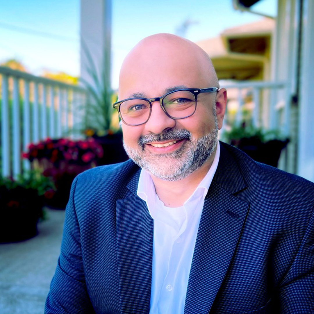 A smiling man with glasses and a beard, wearing a suit, sitting on a porch with plants in the background.