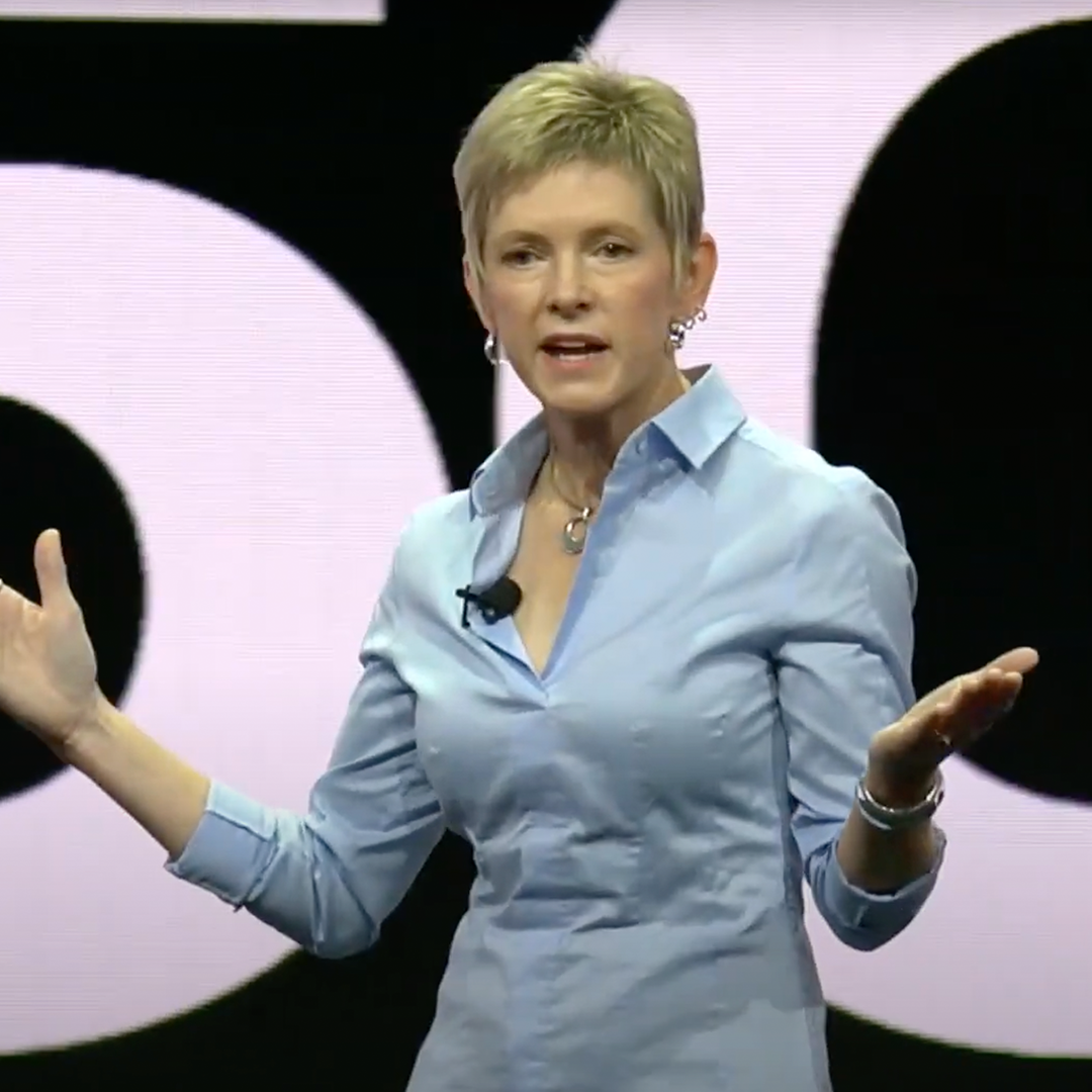 A woman in a blue shirt speaking on stage with a microphone and gesturing with her hands.