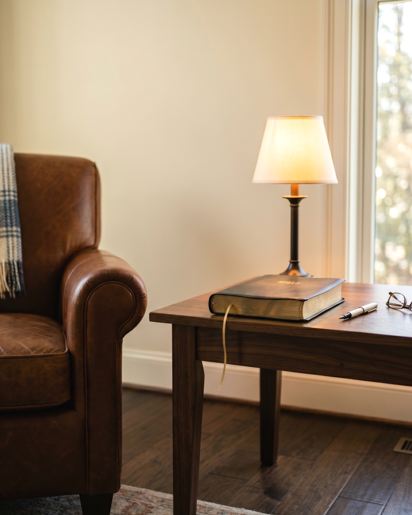 A wooden side table with a closed blue-covered book, a white pen, and glasses on top. A table lamp is nearby, with a brown leather armchair partially visible on the left. There's a window with sunlight coming through, illuminating the room.