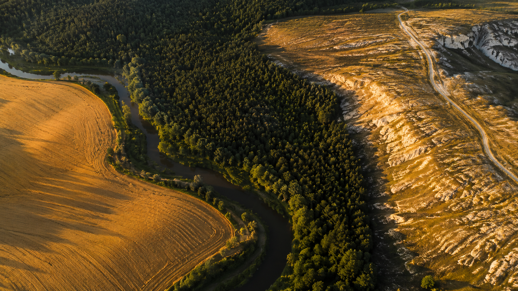 A high-resolution aerial drone photograph showing a natural patchwork landscape divided by organic boundaries. A winding river separates a golden wheat field from dense dark forest, while a sunlit rocky plateau with a dirt road occupies the opposite.