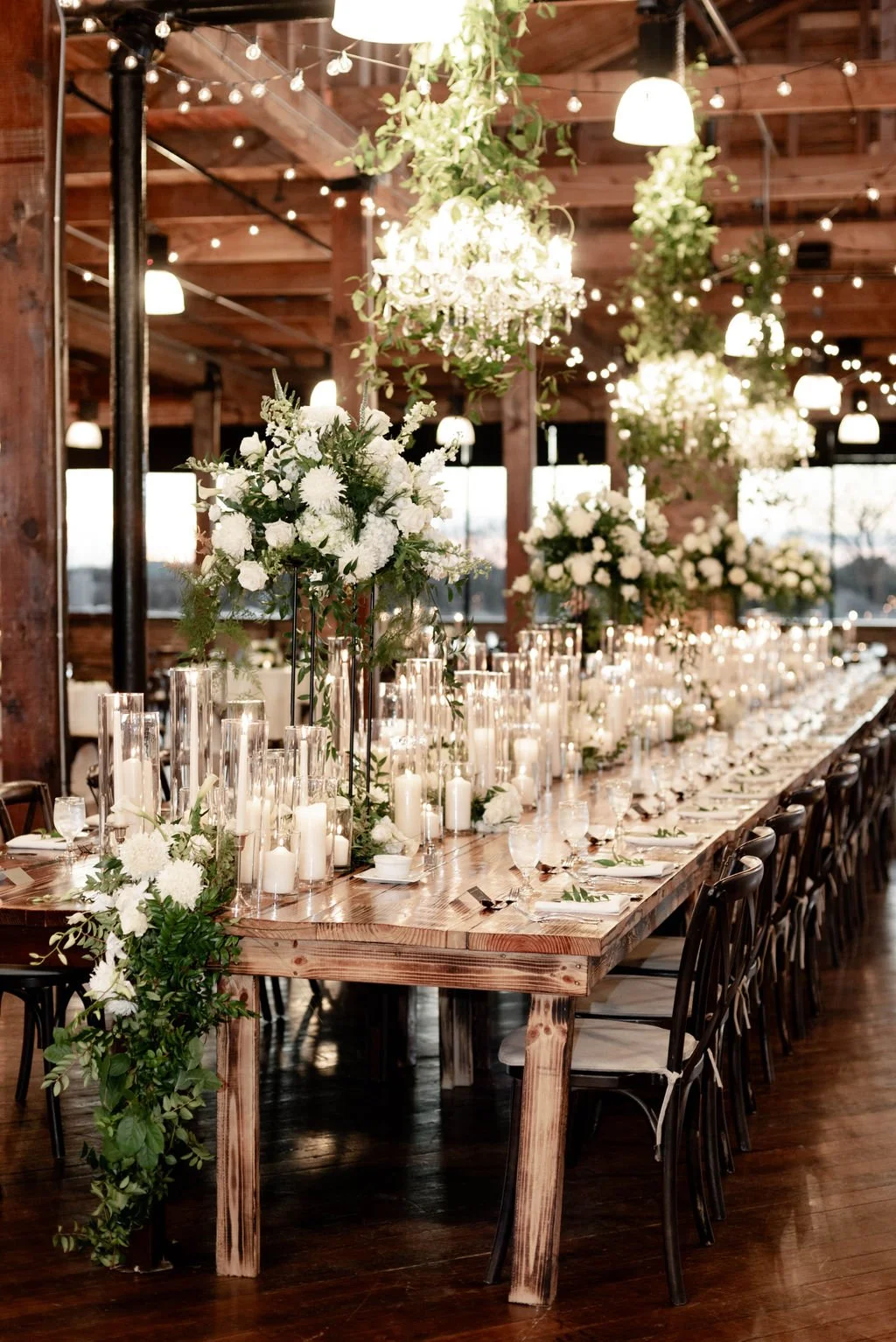 Elegant wedding reception table decorated with white flowers, candles, and greenery inside a rustic wooden venue with hanging chandeliers and string lights.