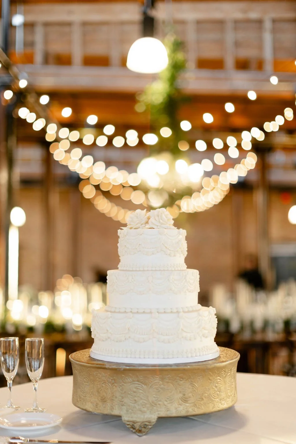 A multi-tiered white wedding cake with ornate piping and decorative flowers on top, sitting on a gold cake stand with two champagne glasses nearby, decorated with string lights in a rustic venue.