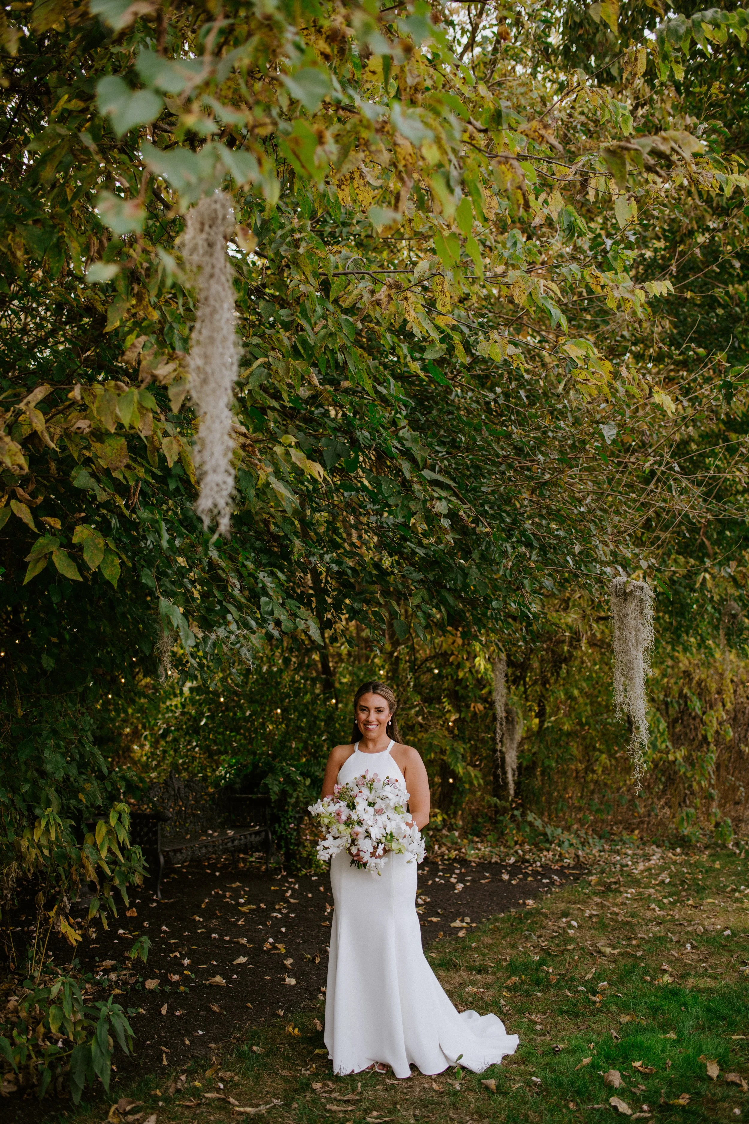 A woman in a white wedding dress standing outdoors in a wooded area with trees and fallen leaves, holding a bouquet of flowers, smiling at the camera.