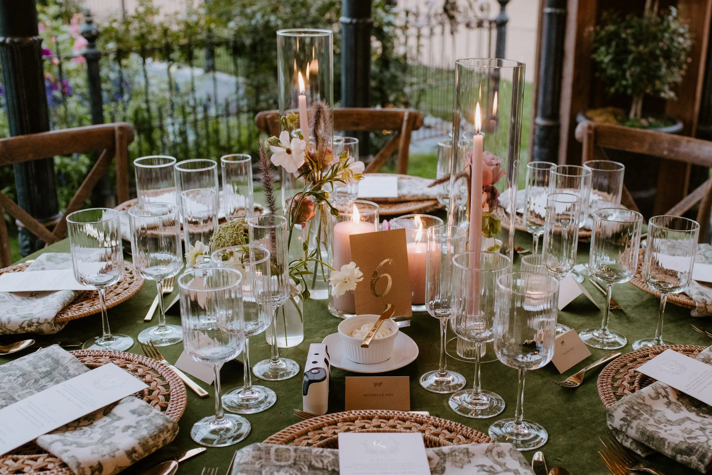 A decorated outdoor dining table set for a celebration with candles, flowers, and glassware, surrounded by wooden chairs and greenery.