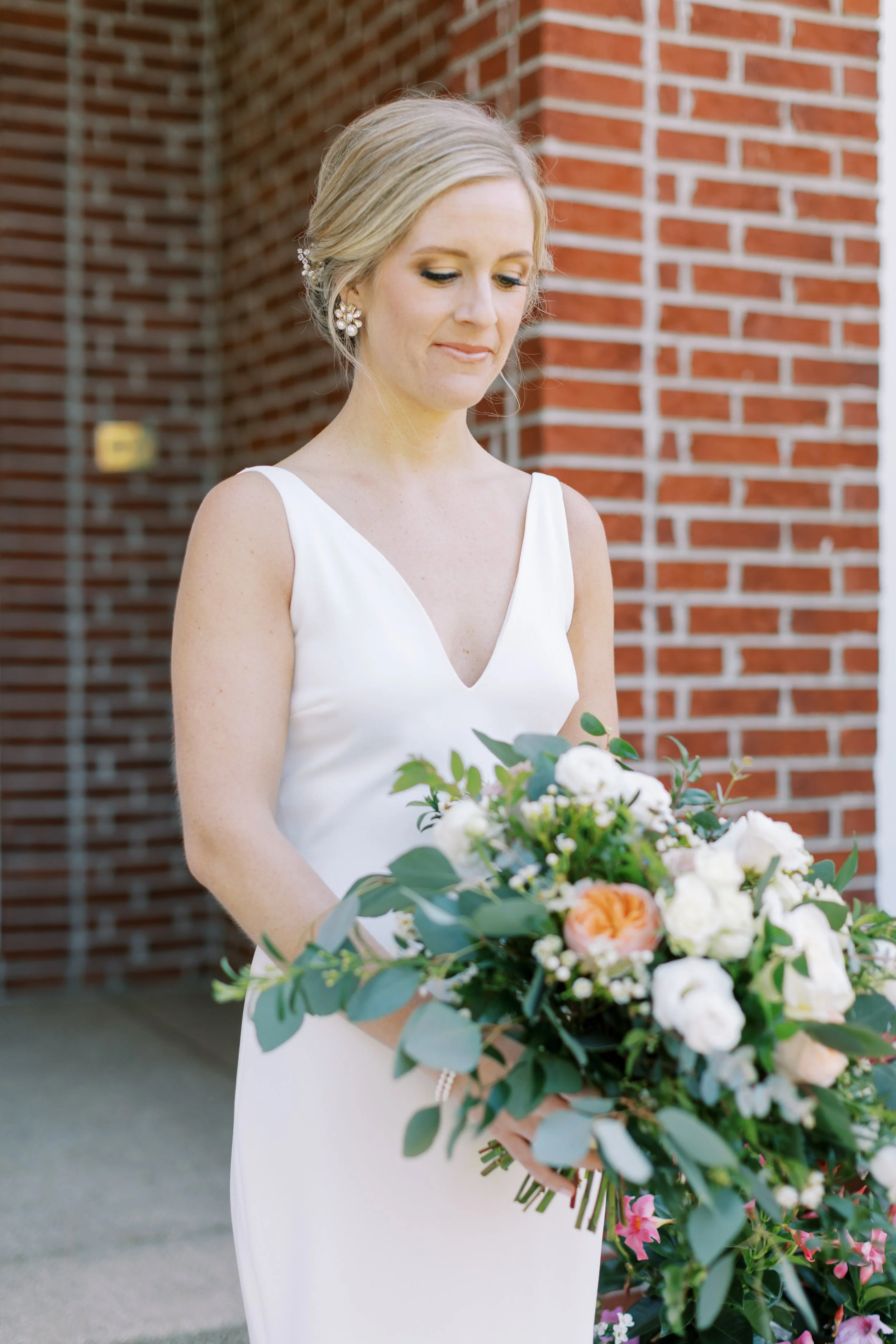 A bride in a white wedding dress holding a large bouquet of flowers, standing outside in front of a red brick wall.