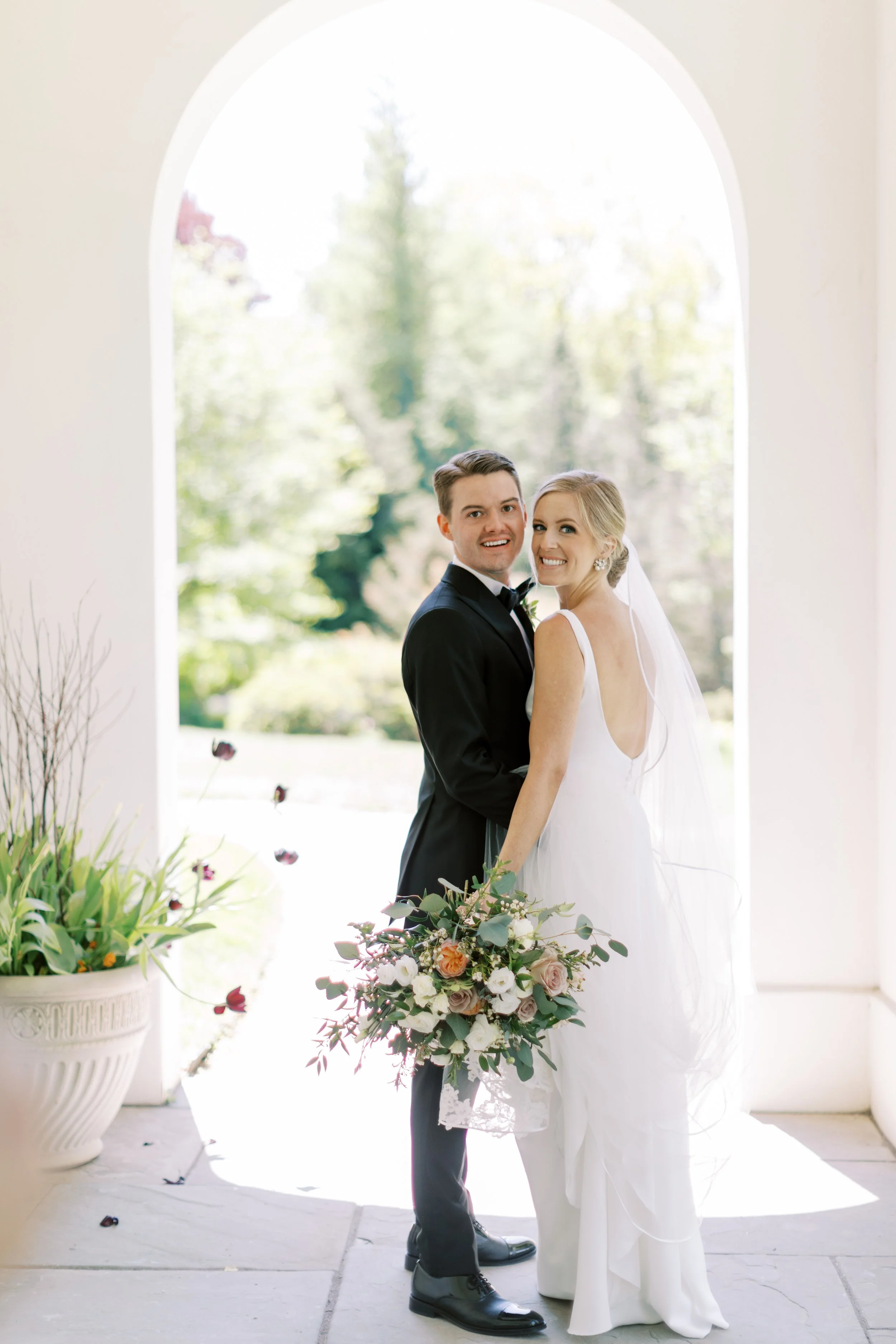 A newlywed couple stands in front of a large arched window with bright natural light shining through, and trees visible outside. The bride wears a white wedding dress with a veil, and the groom wears a black tuxedo with a bowtie. They are smiling and