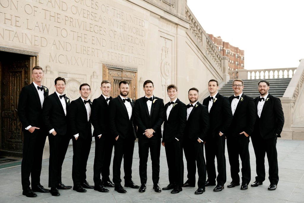 A group of eleven men dressed in black tuxedos with bow ties posing outdoors in front of a large stone building with engraved text and stairs.