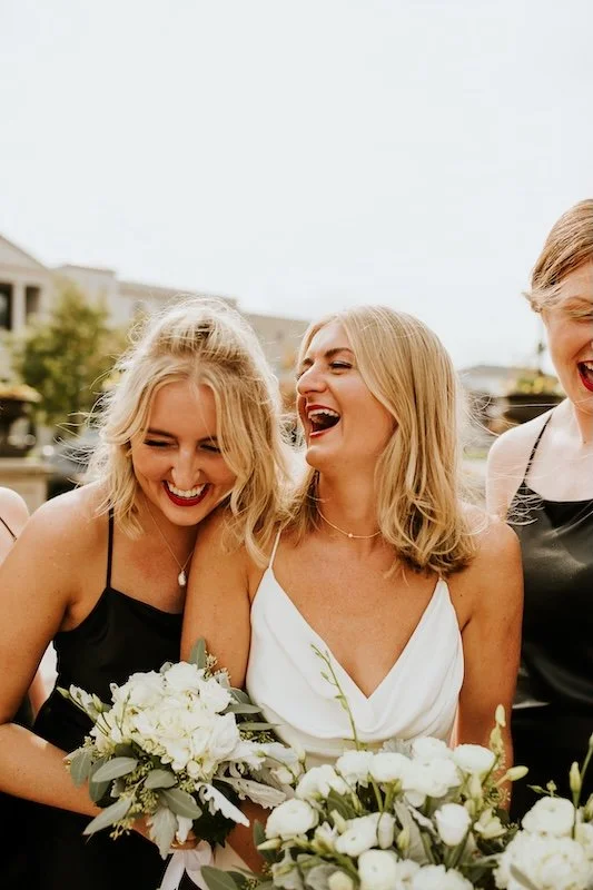 Three women smiling and laughing, holding bouquets of white flowers, at a wedding celebration outdoors.