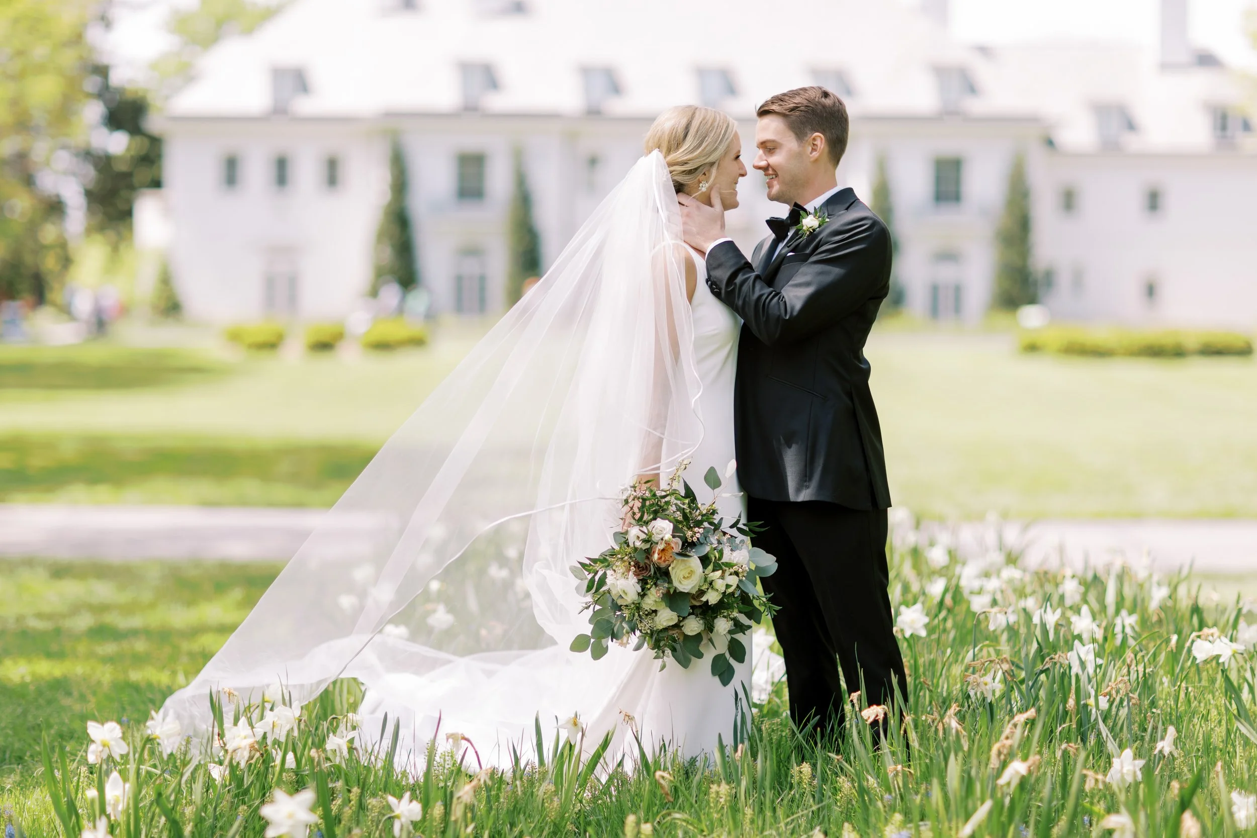 A bride and groom in wedding attire standing close together outdoors, holding a bouquet of flowers, with a large white building in the background and flowers on the grass in the foreground.