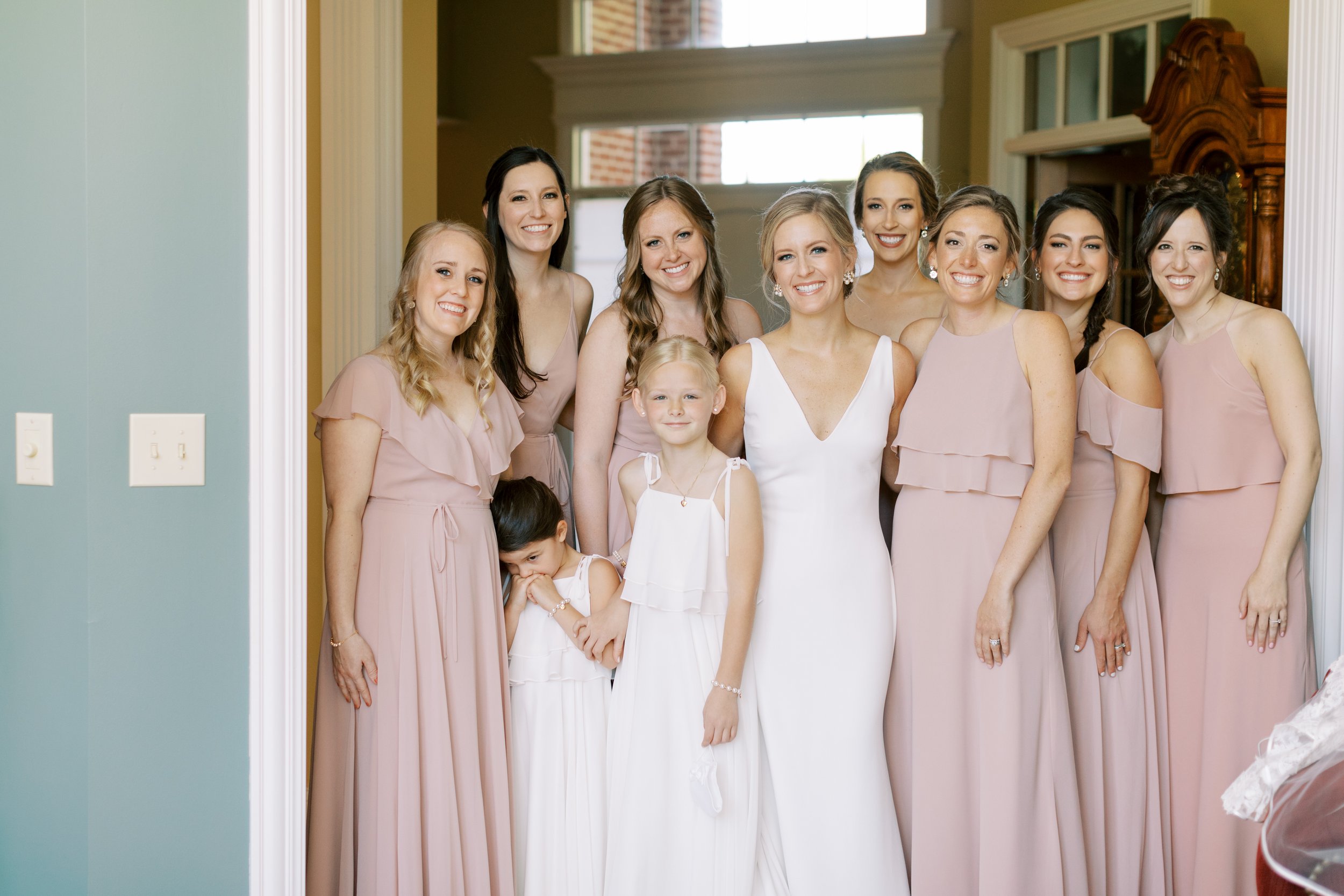 A group of women in pink dresses and two young girls in white dresses smiling for a photo inside a home with large windows.