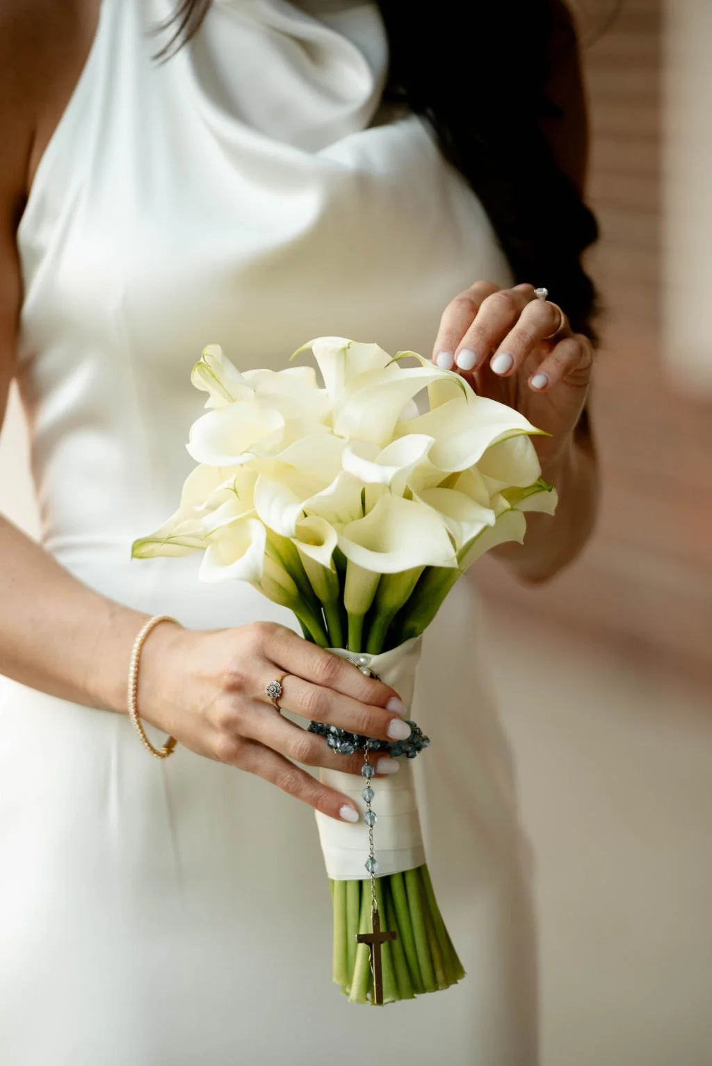 A bride holding a bouquet of white calla lilies, with her hands showing a wedding ring and a bracelet. She is wearing a white dress, and the background appears to be indoors.