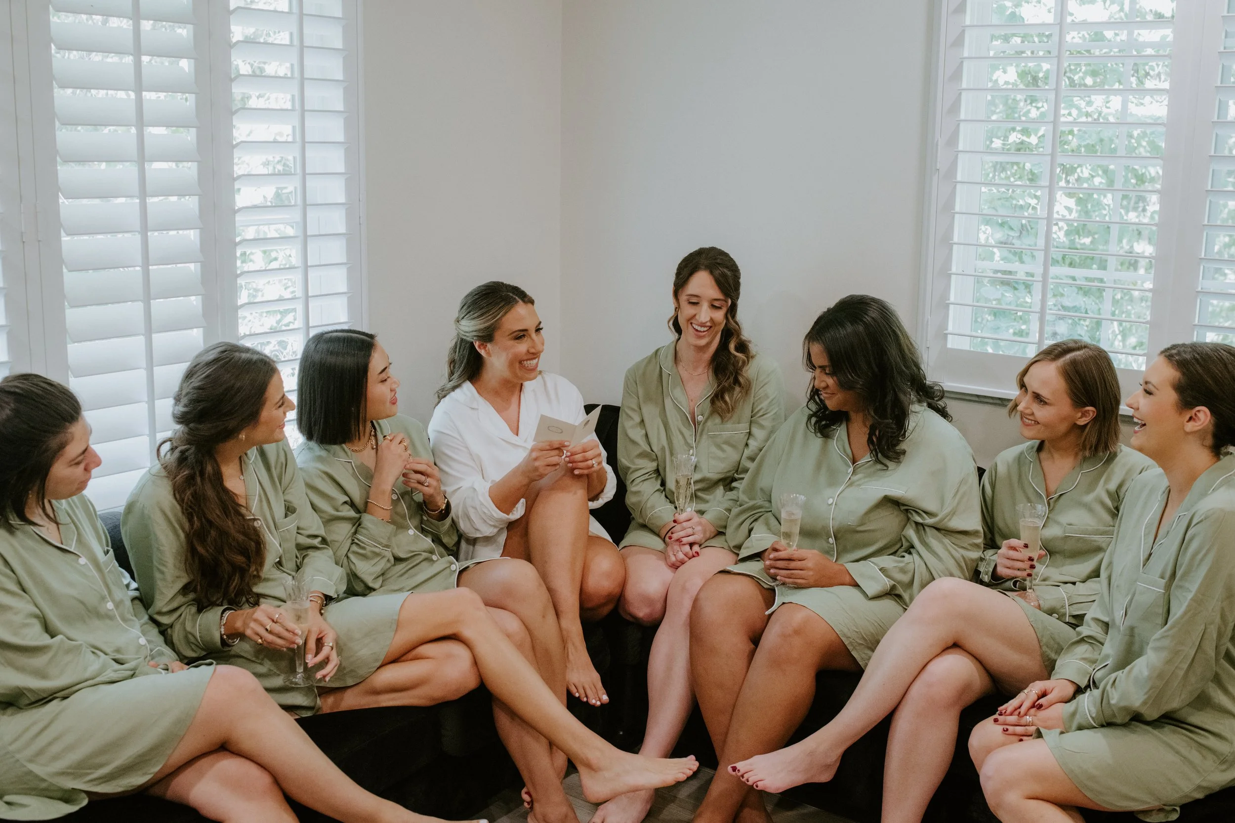 A group of women sitting closely together in a cozy room with large windows and white shutters, some wearing matching green pajamas, while one woman in a different outfit holds a card and drinks champagne, all appear to be enjoying a social gathering