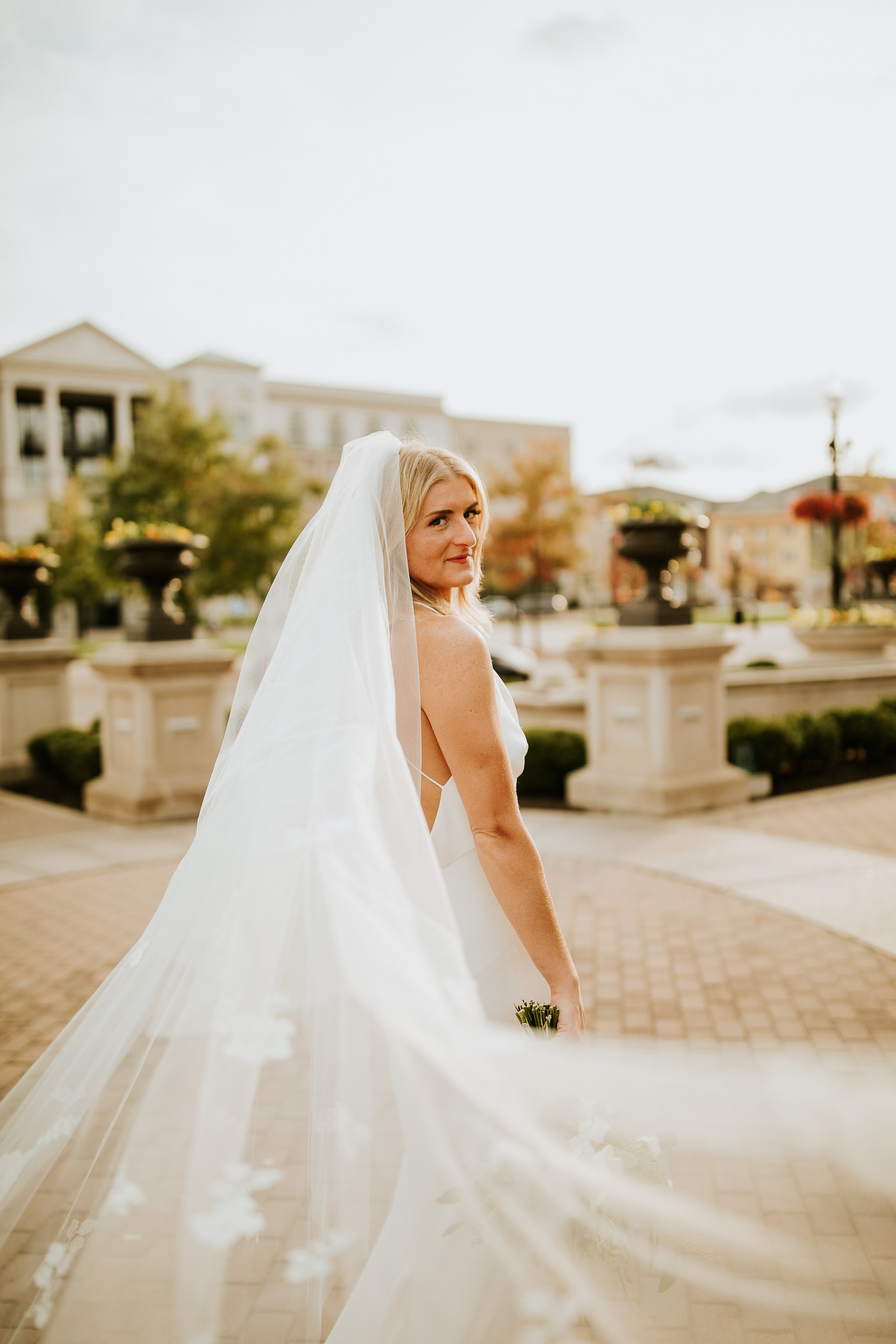 A bride with blonde hair wearing a white wedding dress and veil stands outdoors on a paved area with buildings, trees, and flower pots in the background.