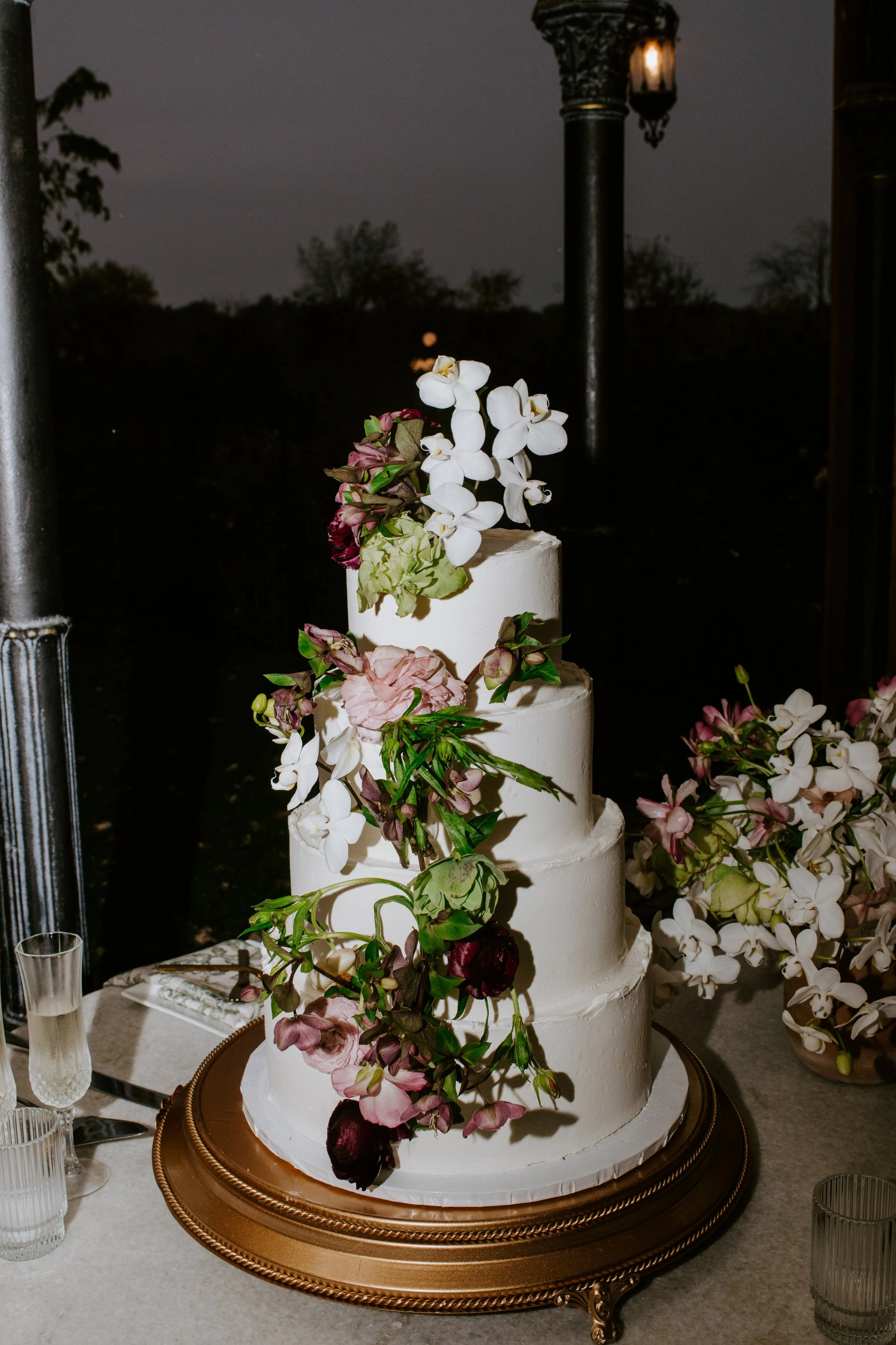 A four-tiered white wedding cake decorated with cascading pink, white, and dark red flowers and greenery, displayed on a decorative gold cake stand at a wedding reception.