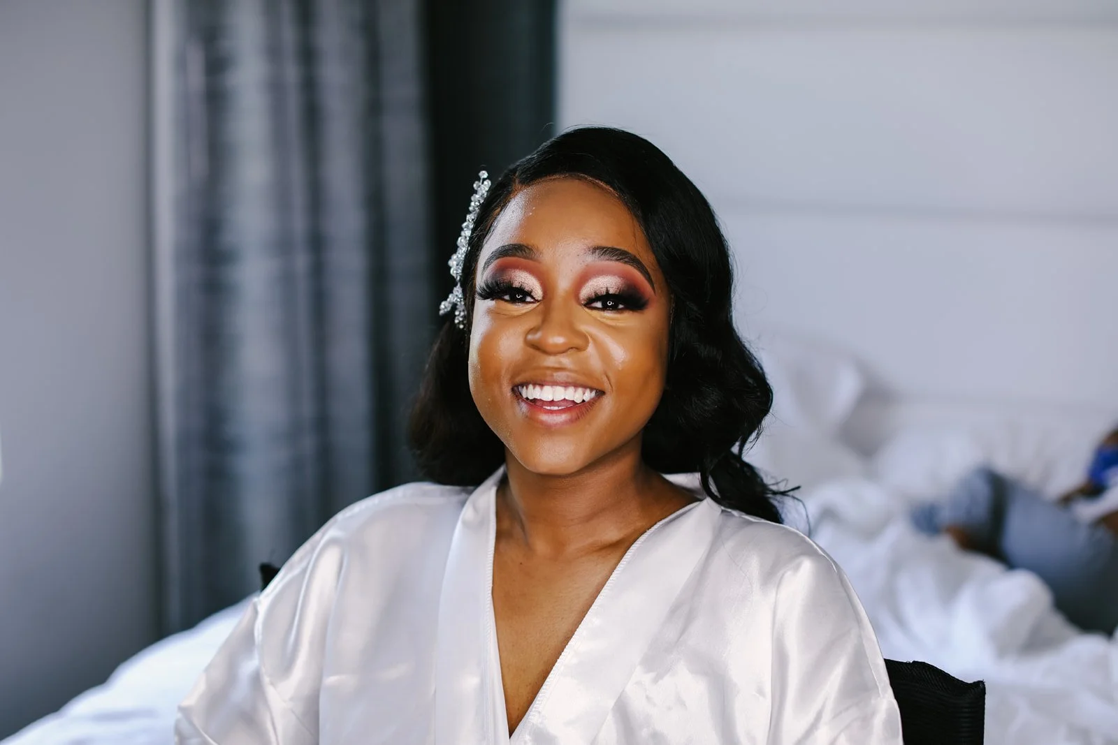 A smiling woman with dark hair styled in waves, wearing makeup and a white satin robe, sitting indoors with a bed in the background.
