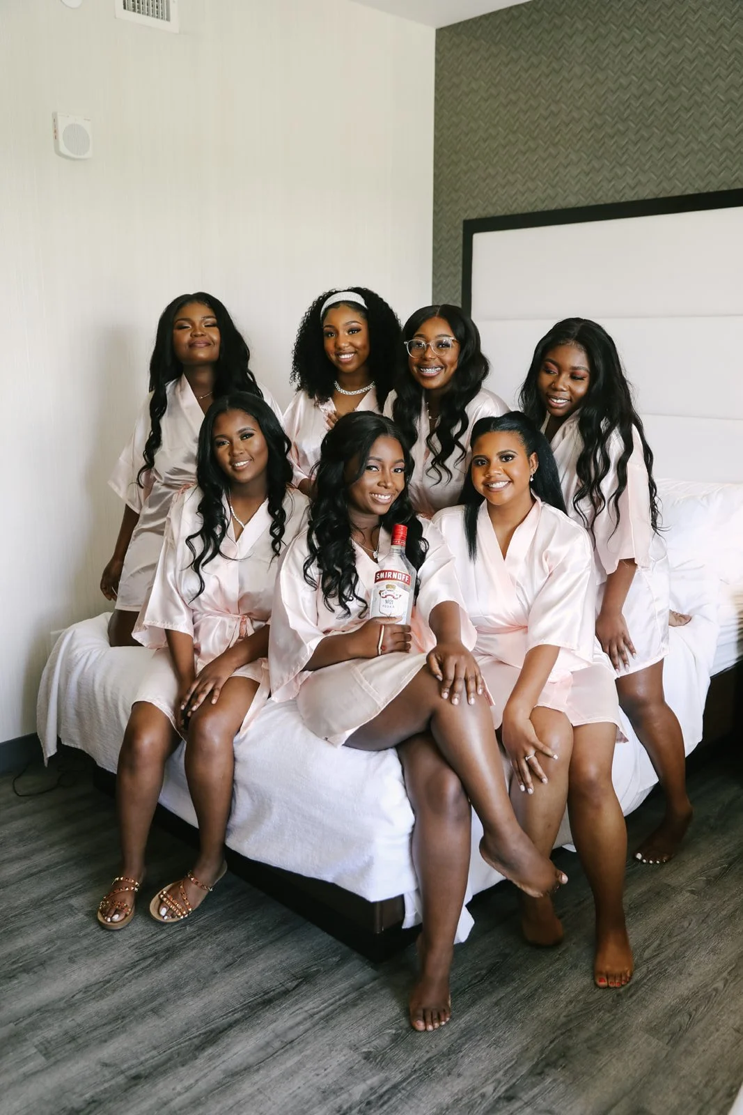 A group of eight women in matching pink satin robes sitting and standing on a bed in a modern hotel room, smiling, with one holding a small bottle of Smirnoff vodka.