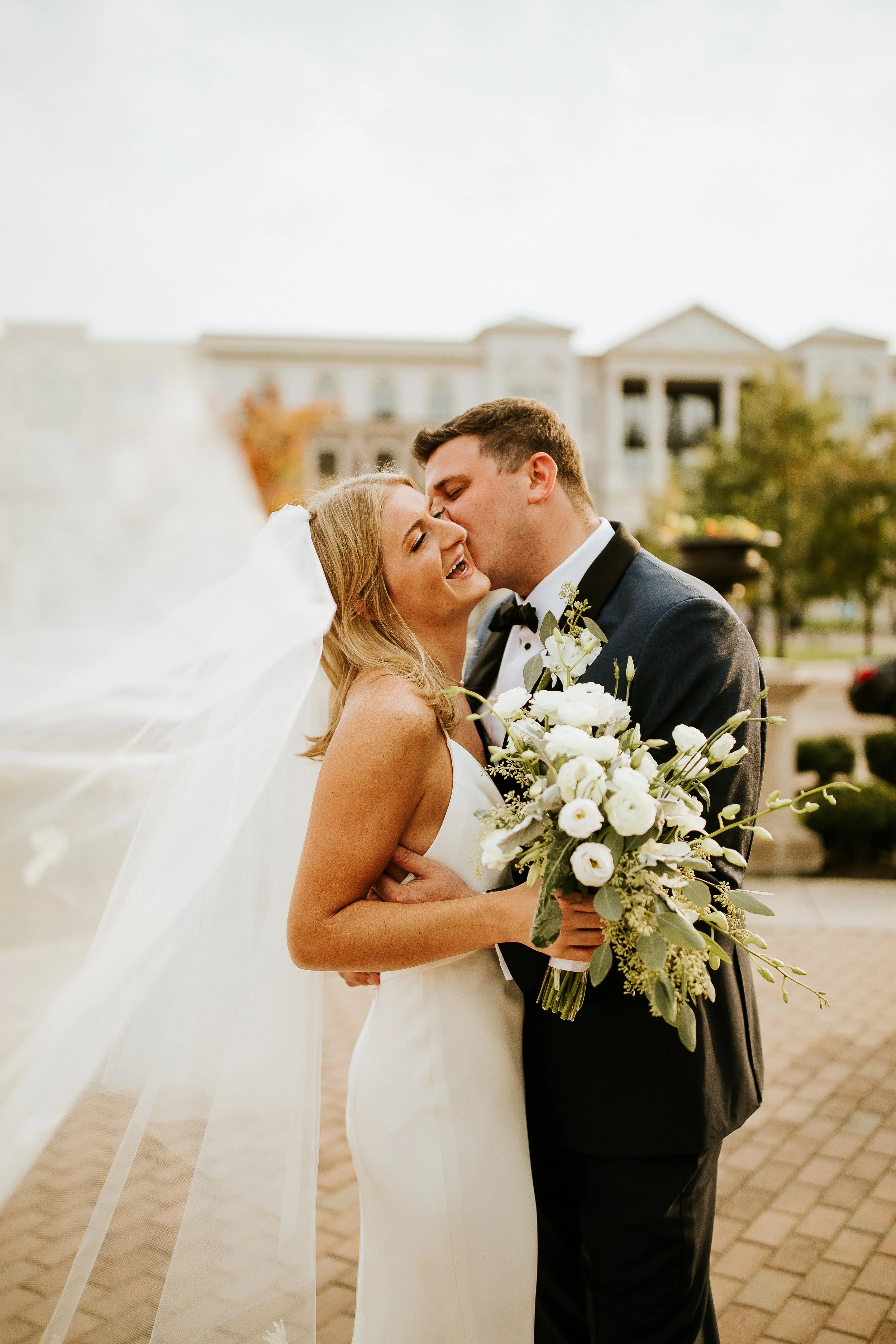 A bride and groom smiling and kissing outdoors, with the bride holding a large bouquet of white flowers, and the groom in a black tuxedo with a boutonniere, in front of a historic building with columns.
