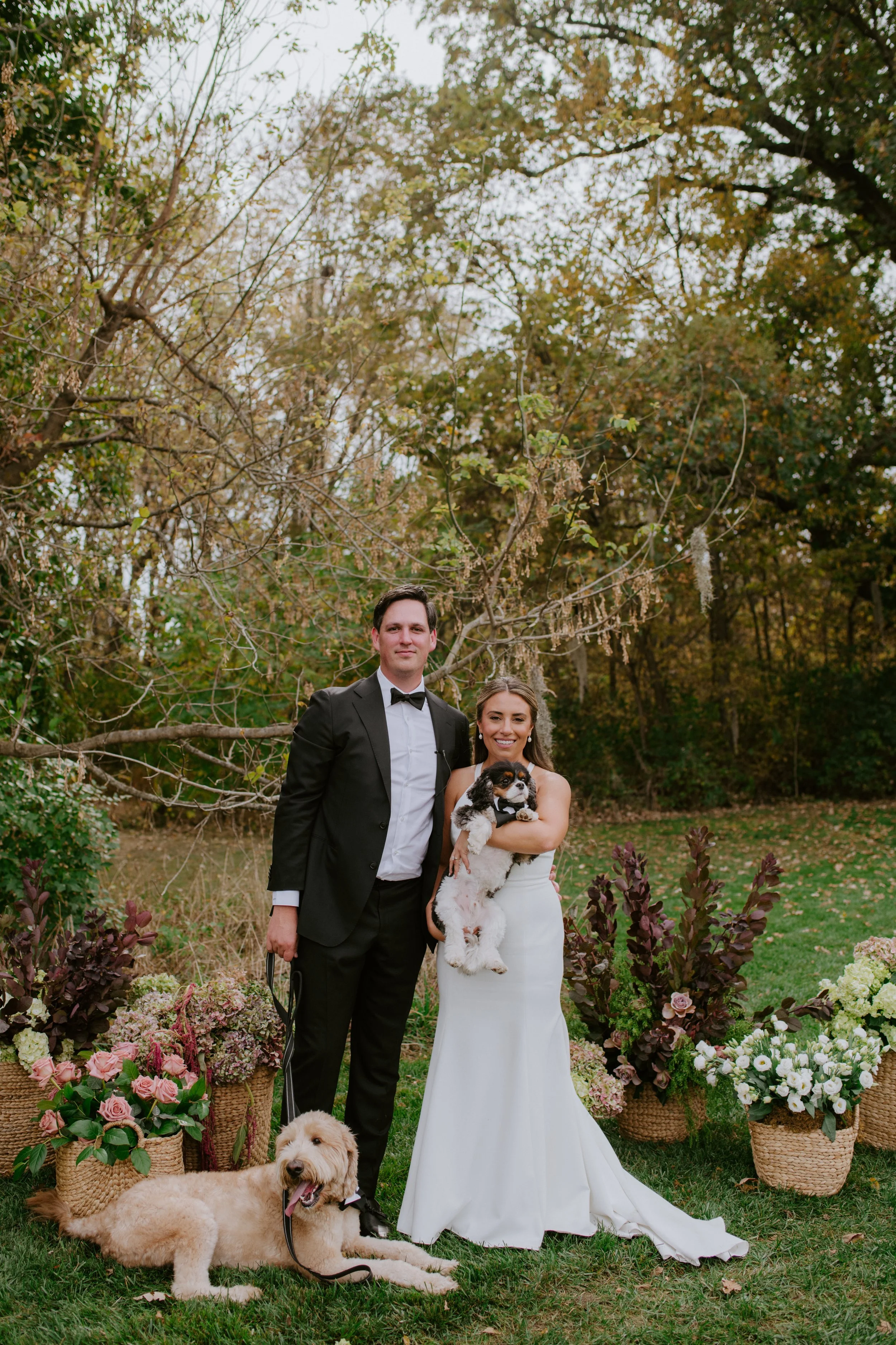 A smiling bride in a white wedding dress holding a black and white puppy, standing beside a groom in a black tuxedo, with a golden retriever lying on the grass, surrounded by baskets of flowers in an outdoor garden setting.