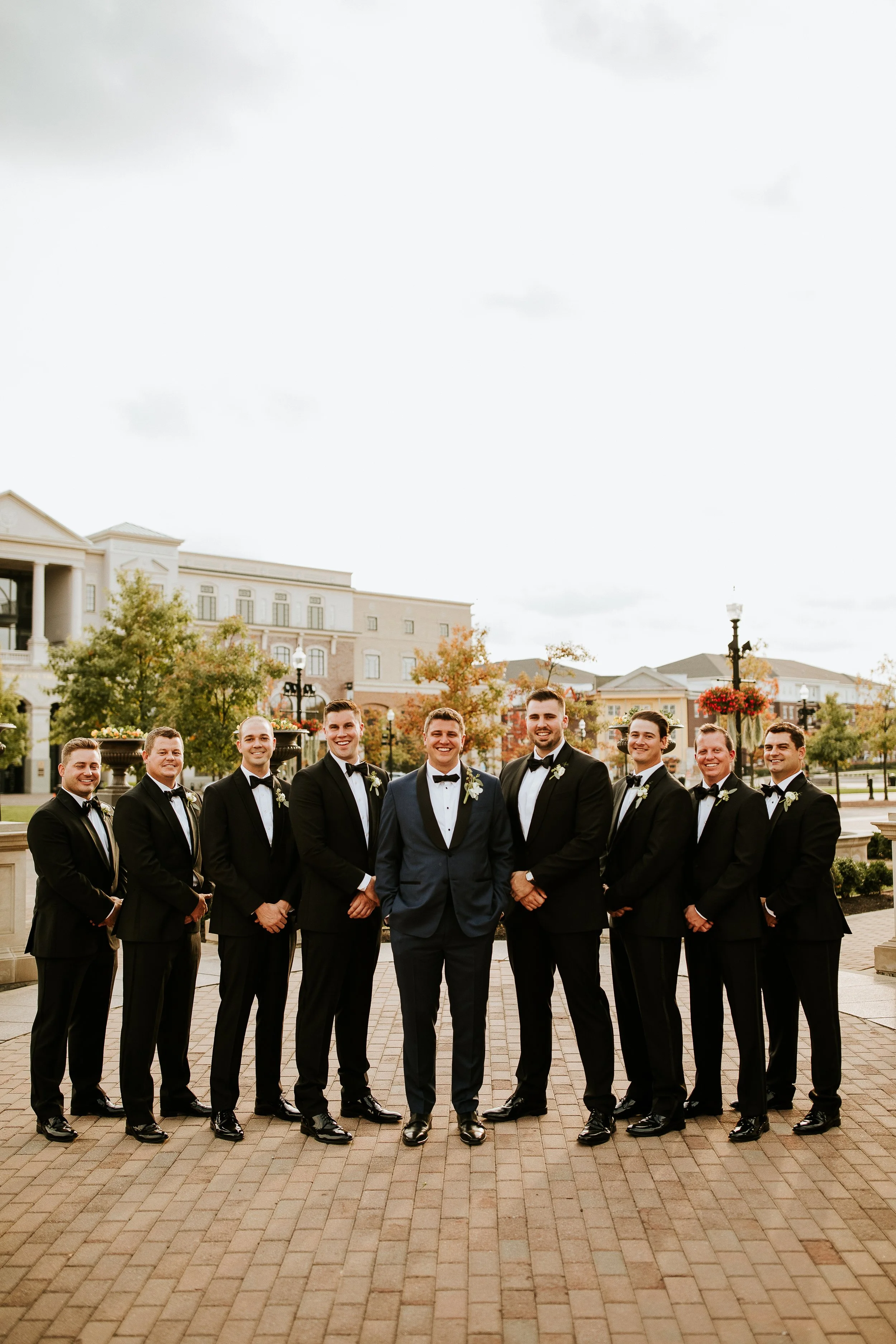 A group of eleven men in tuxedos standing outdoors in front of a cityscape with brick buildings, trees, and street lamps on a paved area. The men are smiling and standing with their hands clasped in front or at their sides.
