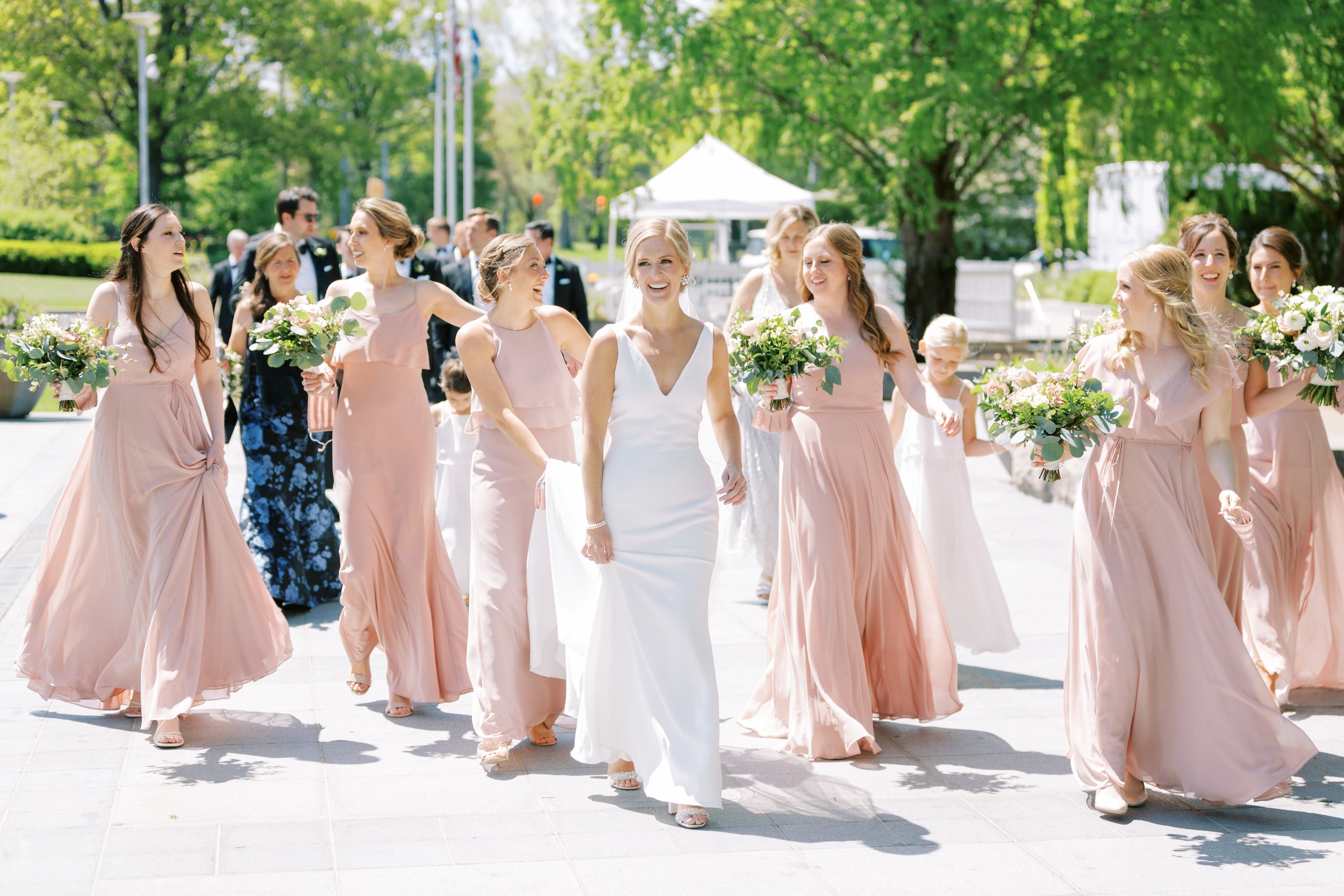 A group of women, dressed in pink and white, walking outdoors at a wedding ceremony while holding bouquets of white and green flowers.