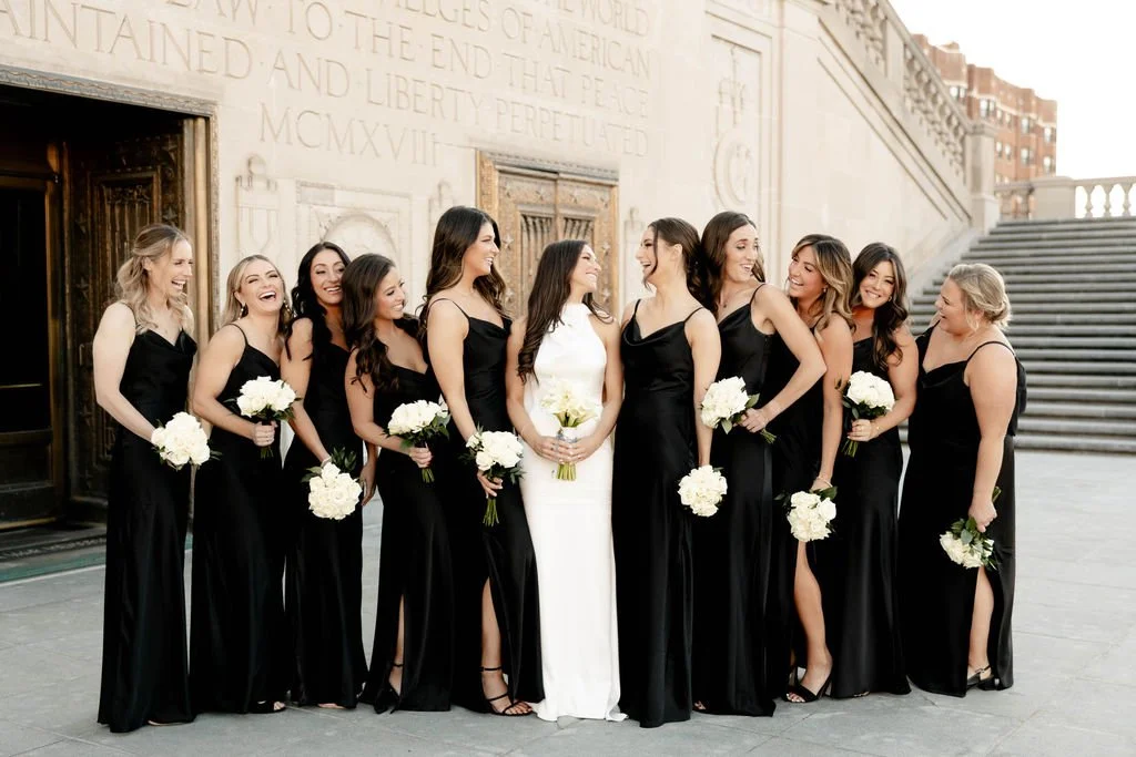 Group of women in black dresses and one woman in a white dress, holding white bouquets, standing outdoors near stairs, smiling and laughing together.