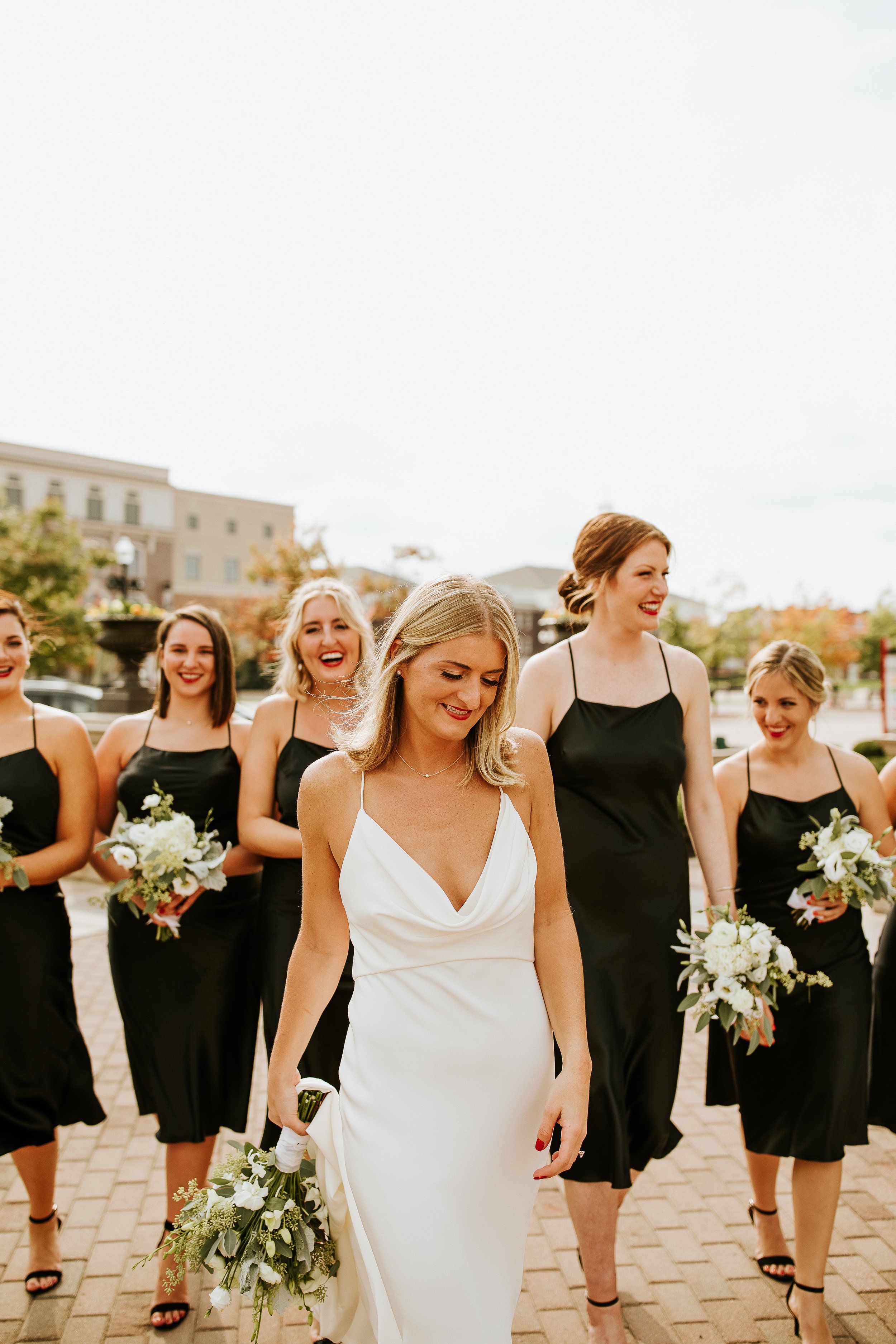 A group of women, including a bride in a white wedding dress and bridesmaids in black dresses, walking outdoors on a brick pavement, holding bouquets of white flowers, with a building and overcast sky in the background.