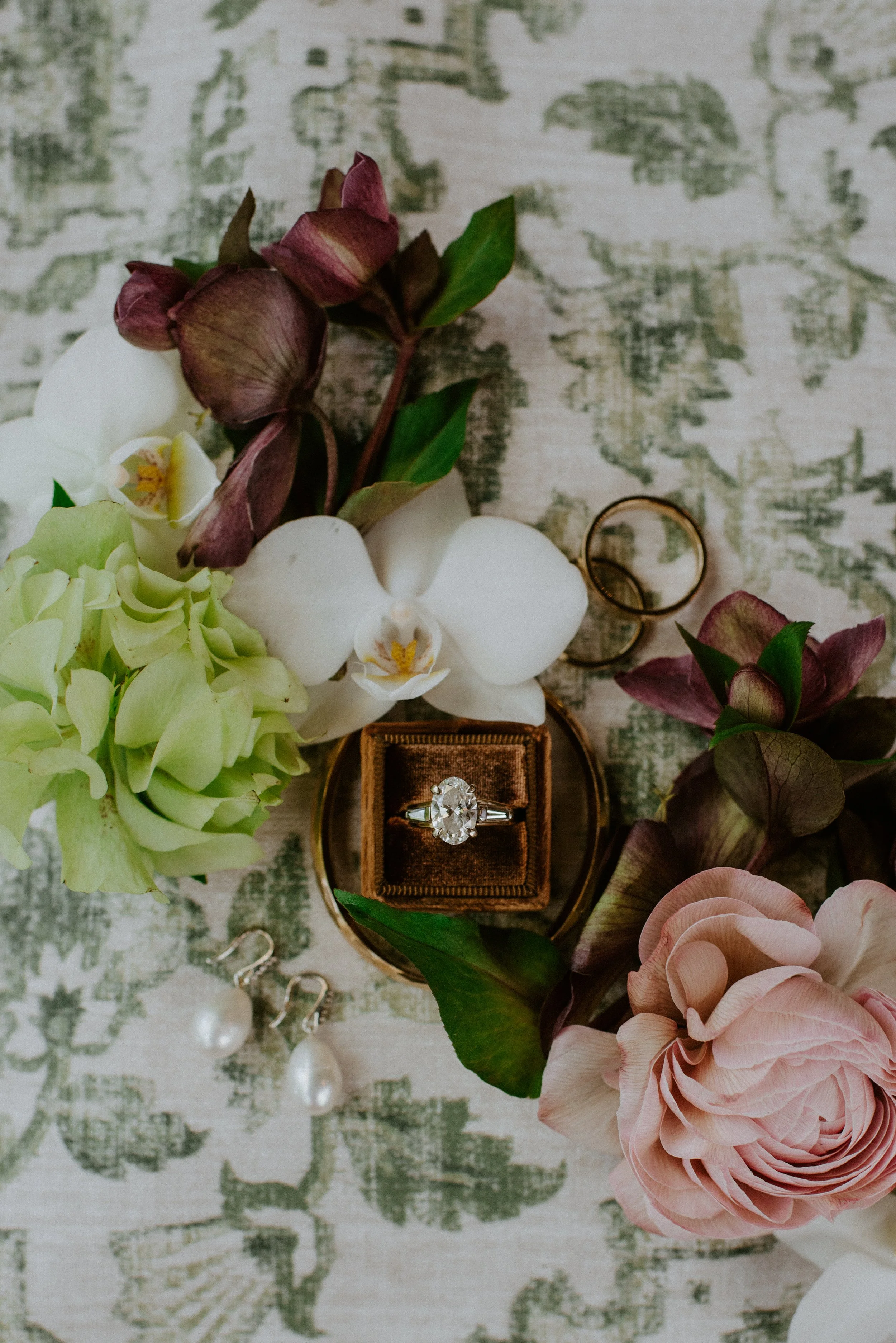 An engagement ring with a large central diamond in a velvet box, surrounded by pink, white, and green flowers, with a pair of wedding rings nearby, all on a textured surface.